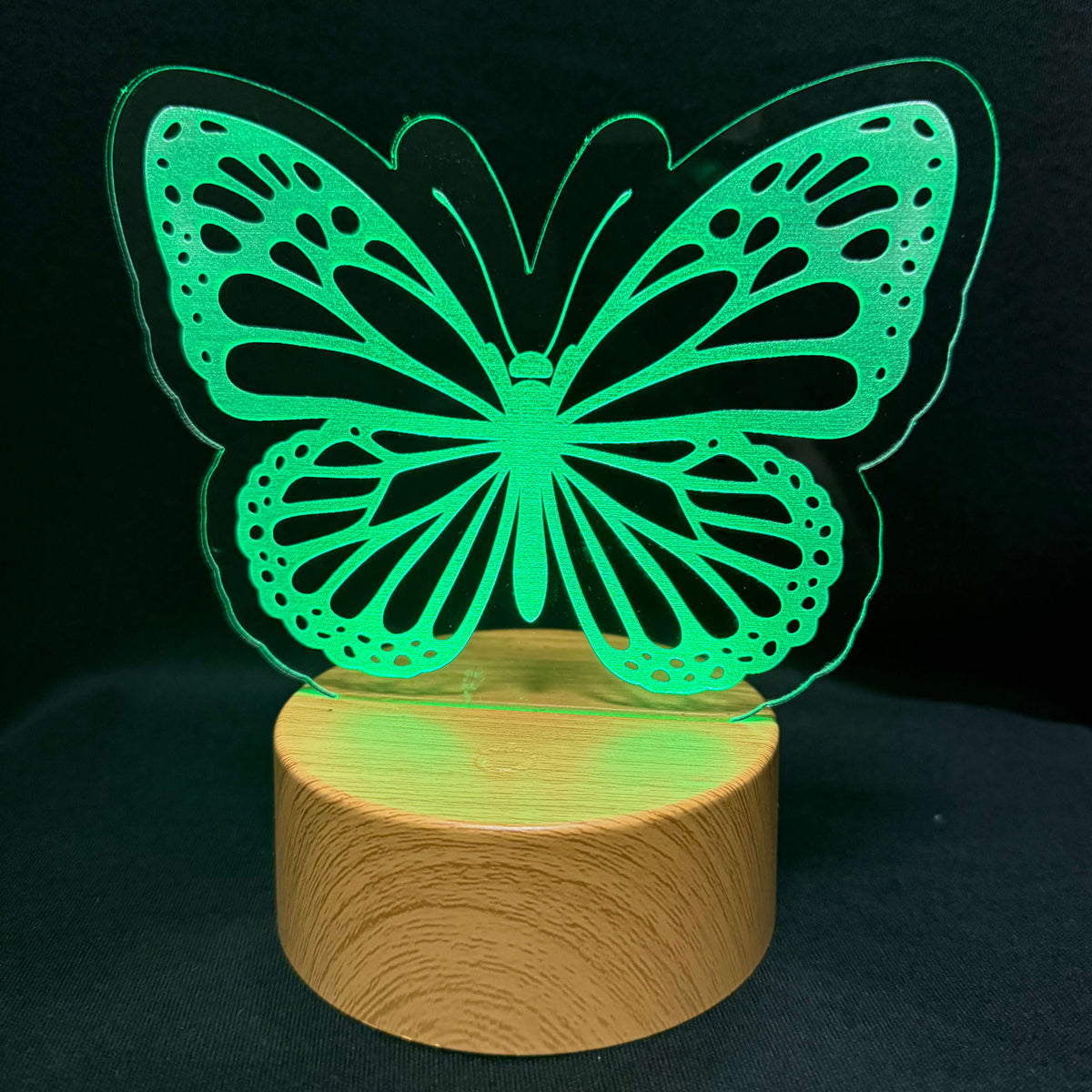 A glowing green butterfly-shaped LED night light on a round wooden base, set against a dark black background.