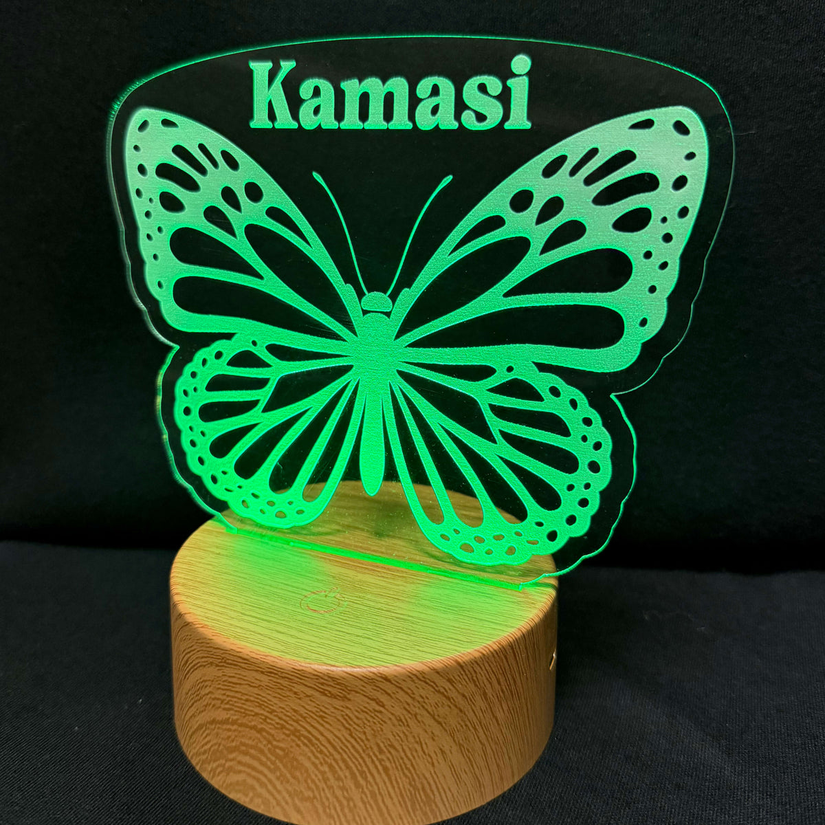 A personalized acrylic LED night light featuring a glowing green butterfly design and the name 'Kamasi' in matching green text, mounted on a round wooden base against a black background.