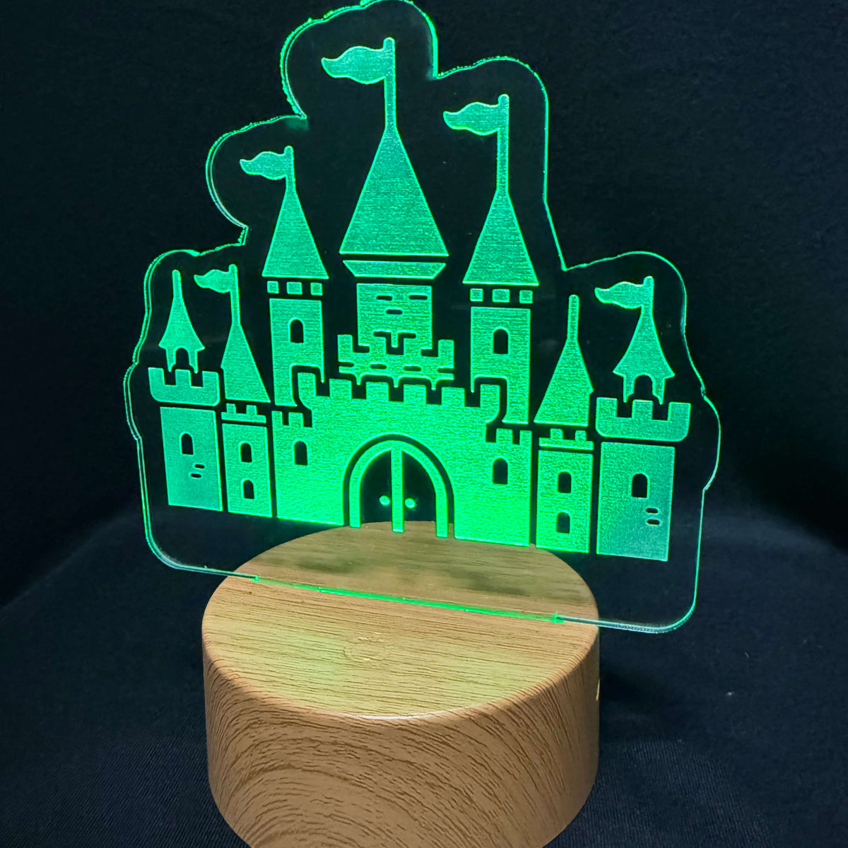 A wooden cylindrical night light base with a clear acrylic castle-shaped panel glowing green, set against a dark black background.