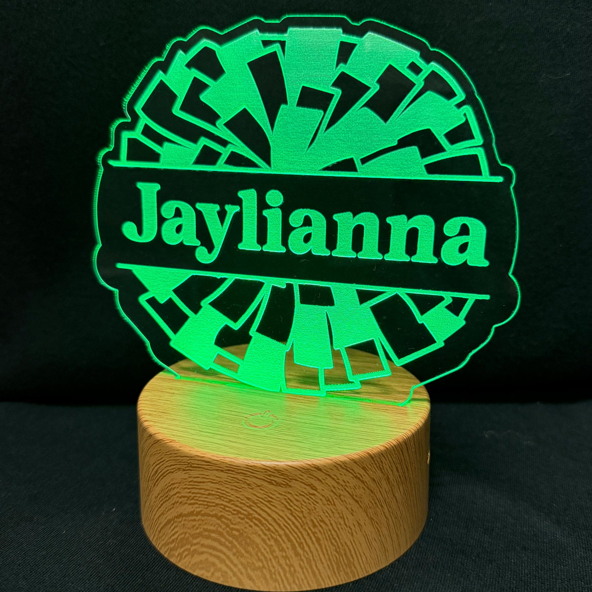 A personalized LED night light with a circular laser-cut design spelling 'Jaylianna' in green, illuminated to glow against a black background, mounted on a round wooden base.