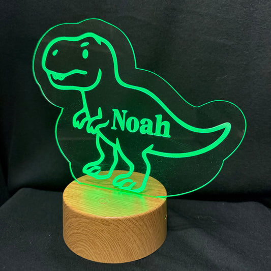 A personalized LED acrylic night light featuring a green glowing outline of a T-Rex dinosaur with the name 'Noah' engraved on its side, mounted on a round wooden base against a black fabric background.