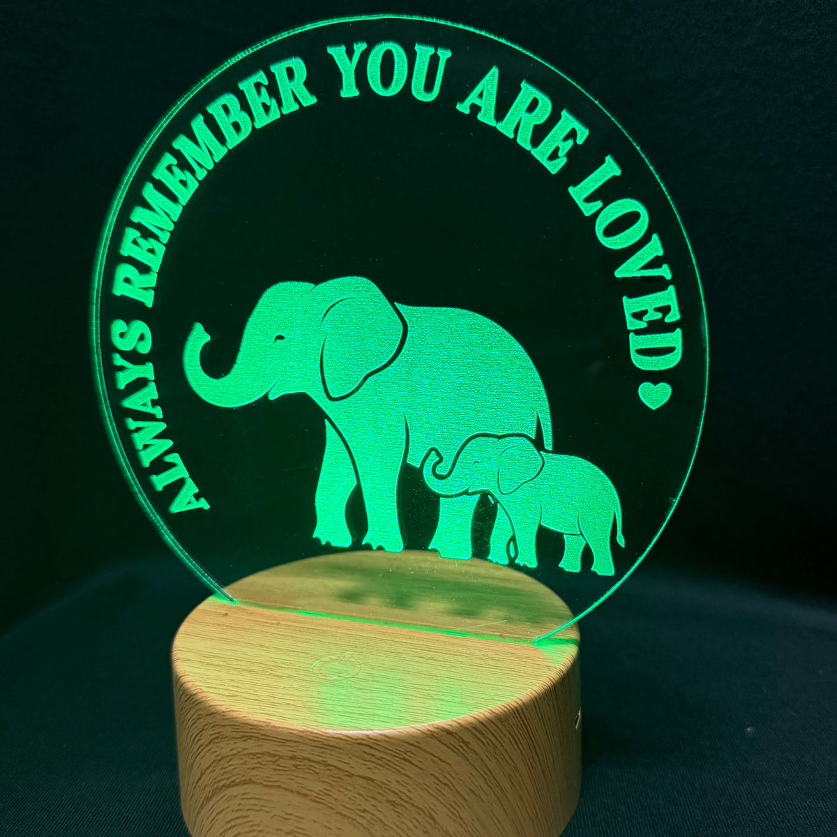 A wooden cylindrical night light base with a clear acrylic lamp panel glowing green. The panel features an elephant and baby elephant silhouette with the text 'ALWAYS REMEMBER YOU ARE LOVED' in a circular layout, set against a dark background.
