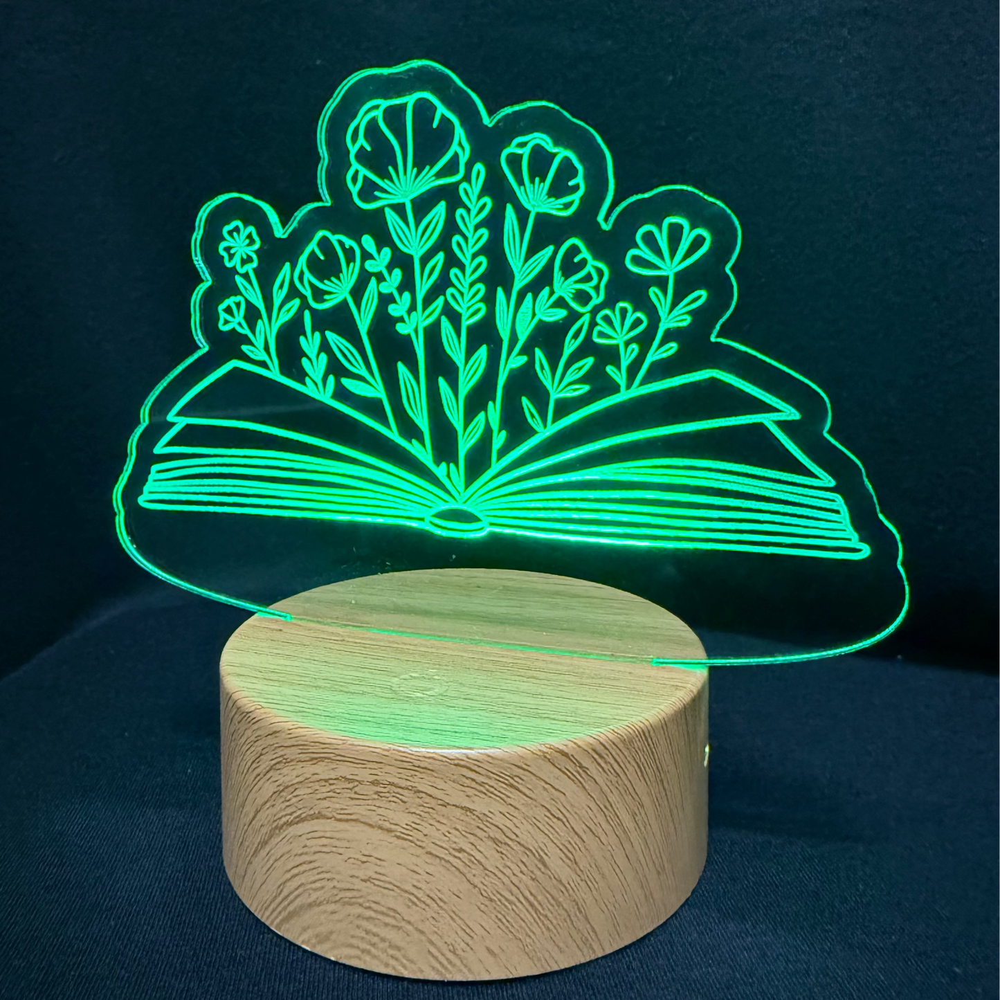 A 3D optical illusion night light featuring an open book with glowing green flowers, resting on a cylindrical wooden base against a dark blue background.