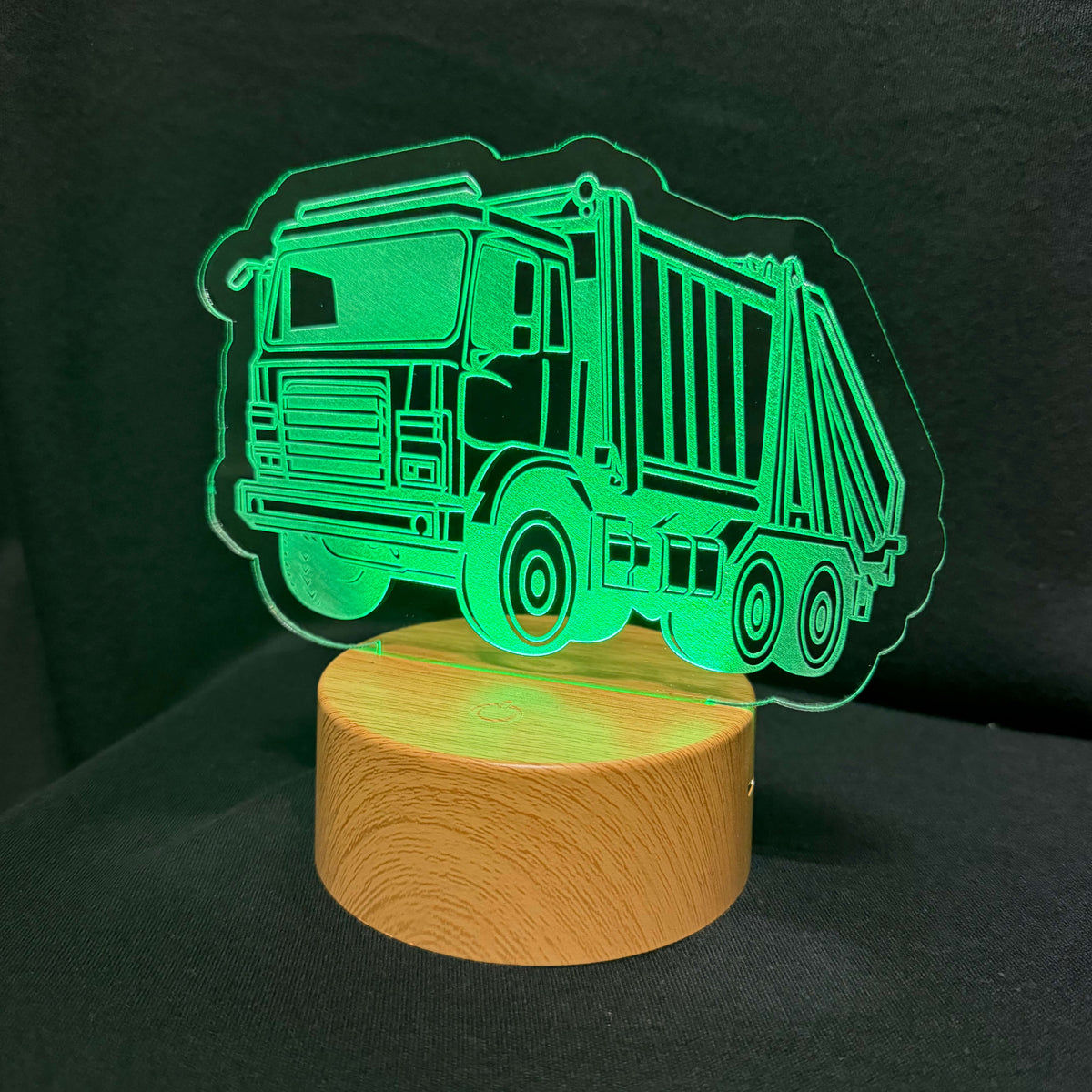 A truck-shaped LED night light with a glowing green laser-cut acrylic design mounted on a round wooden base, photographed against a dark background.