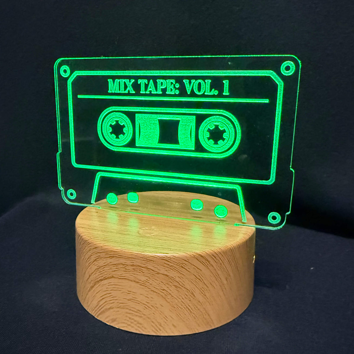 A glowing green cassette tape-shaped LED light on a round wooden base, set against a dark black background. The cassette features the text 'MIX TAPE: VOL.