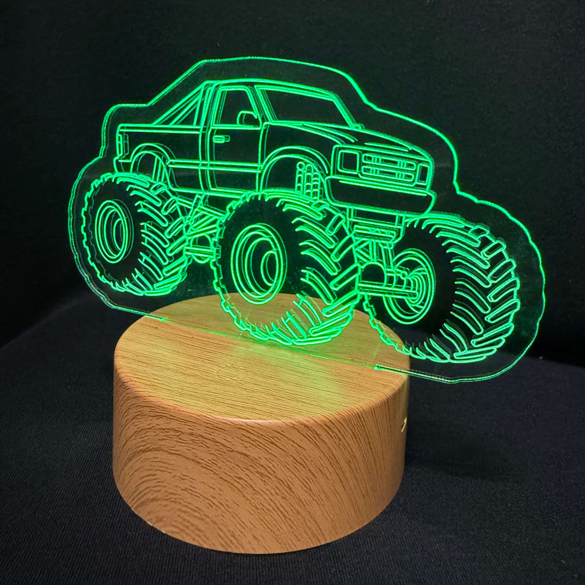 A 3D optical illusion night light featuring a glowing green monster truck outline on a round wooden base, set against a dark black background.