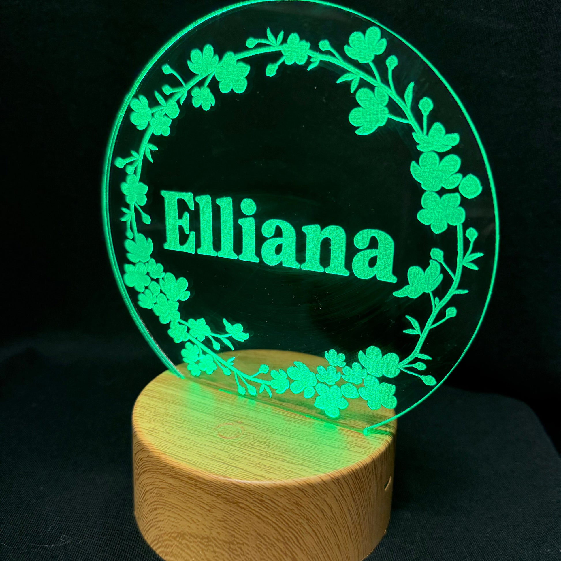 A personalized LED acrylic lamp featuring the name 'Elliana' in bright green text, surrounded by a floral wreath design, mounted on a cylindrical wooden base against a black background.