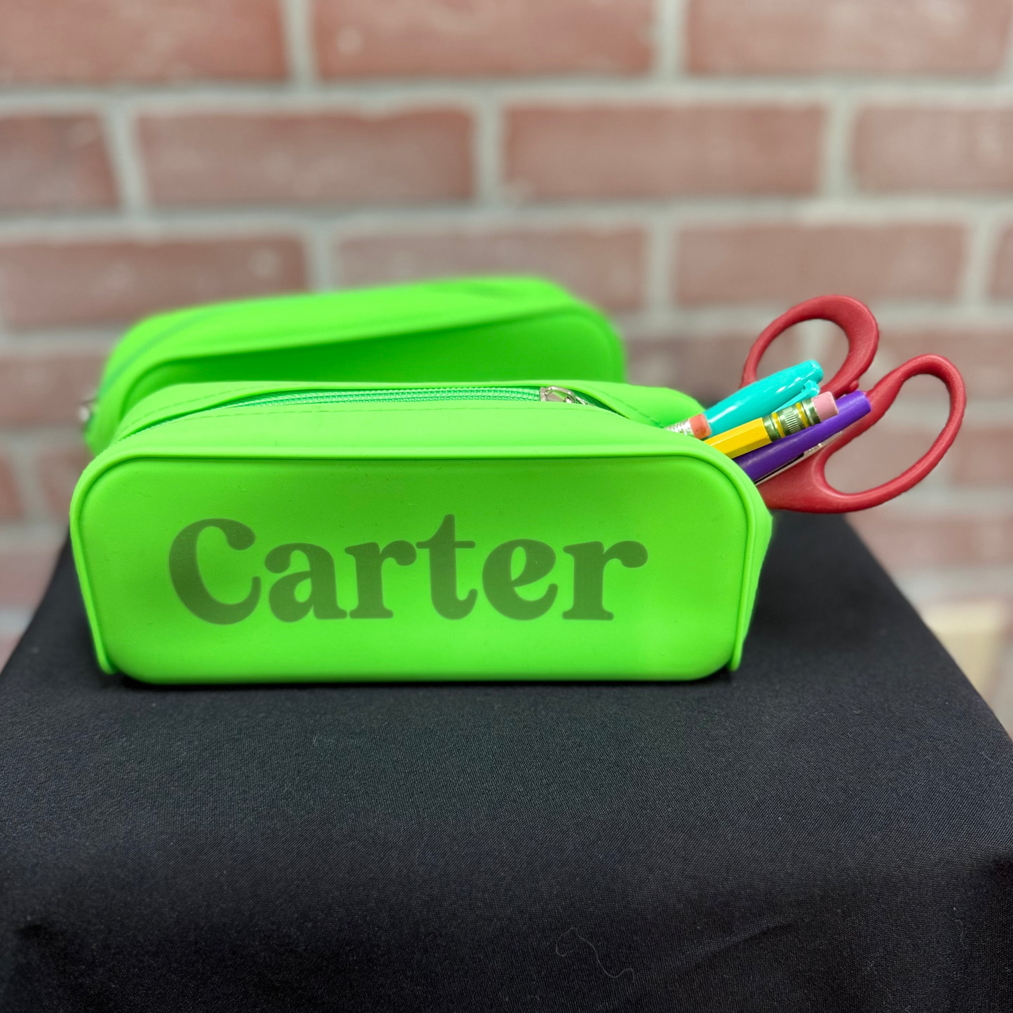 A bright lime green personalized pencil case with the name 'Carter' printed on the front, sitting on a black tablecloth in front of a brick wall. A pair of red scissors and colorful pens are tucked into the zippered pocket.