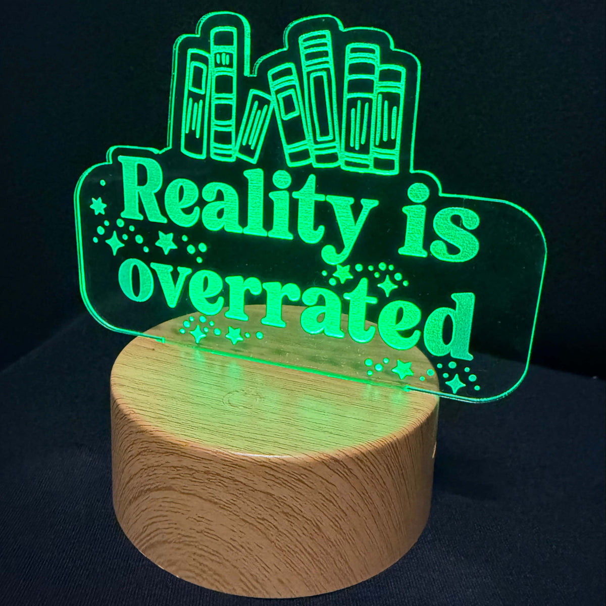 A novelty LED night light with a cylindrical wooden base and a glowing green acrylic sign that reads 'Reality is overrated' with cartoon buildings and stars, shown against a dark background.