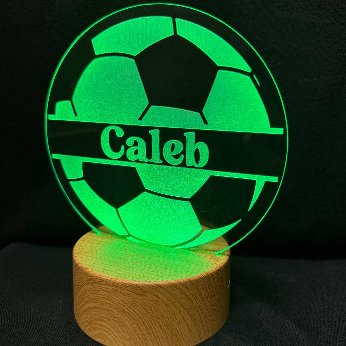 A personalized LED acrylic lamp featuring a soccer ball design with the name 'Caleb' illuminated in green, resting on a round wooden base against a black background.