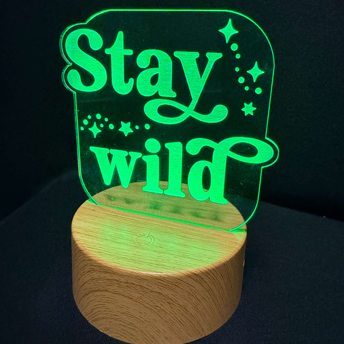 A 3D LED acrylic lamp featuring the phrase 'Stay wild' in glowing green script and star accents, mounted on a cylindrical light wood base against a dark background.