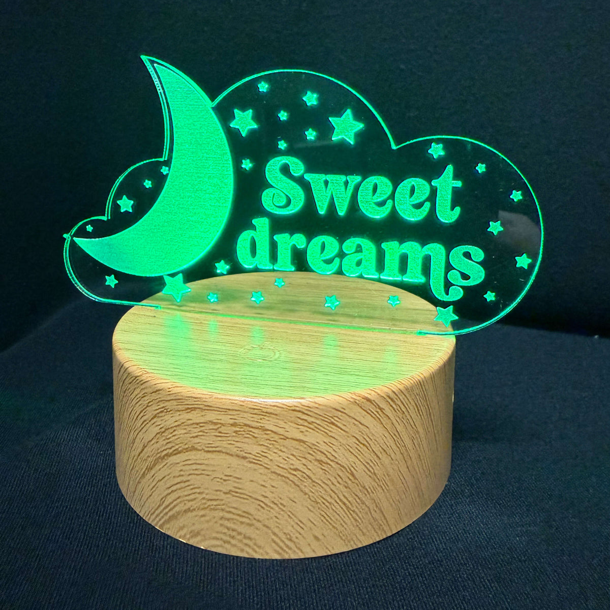A glowing acrylic night light shaped like a cloud with a crescent moon and stars, featuring the words 'Sweet dreams' in green, displayed on a cylindrical wooden base against a dark background.