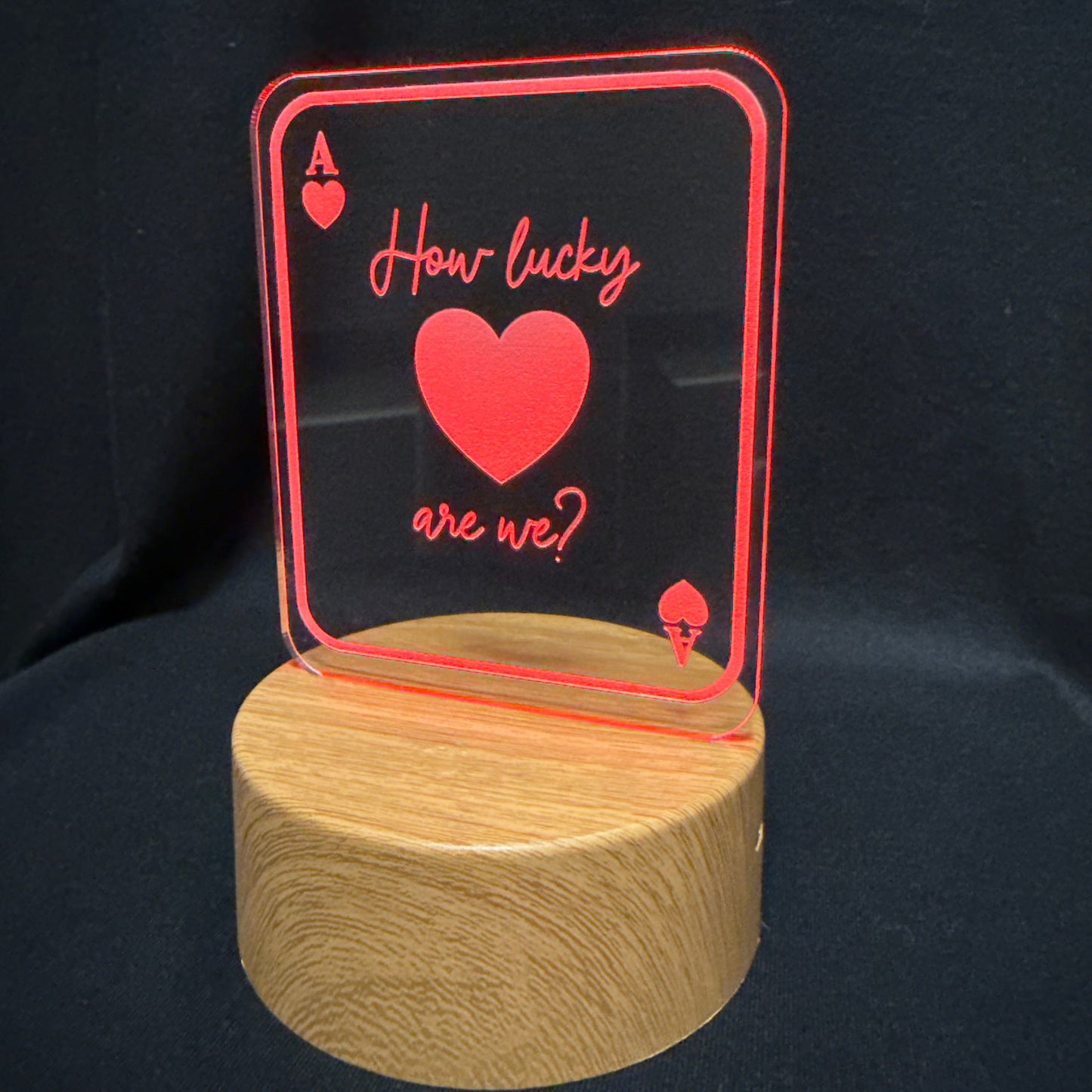 A wooden cylindrical base supporting a clear acrylic sign shaped like an ace of hearts card, with red border lines, a red heart, and the phrase 'How lucky are we?' in red script, set against a black fabric background.