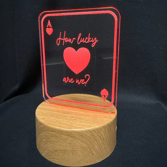 A wooden cylindrical base supporting a clear acrylic sign shaped like an ace of hearts card, with red border lines, a red heart, and the phrase 'How lucky are we?' in red script, set against a black fabric background.