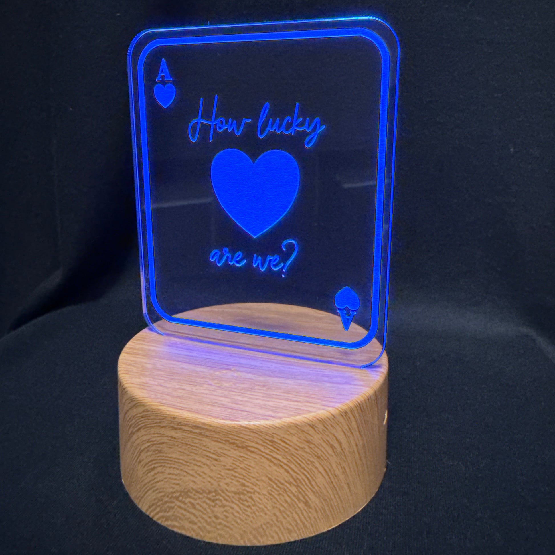 A 3D laser-engraved acrylic LED night light shaped like an Ace of Hearts card, displaying 'How lucky are we?' in glowing blue text and a heart design, mounted on a cylindrical wooden base against a black background.