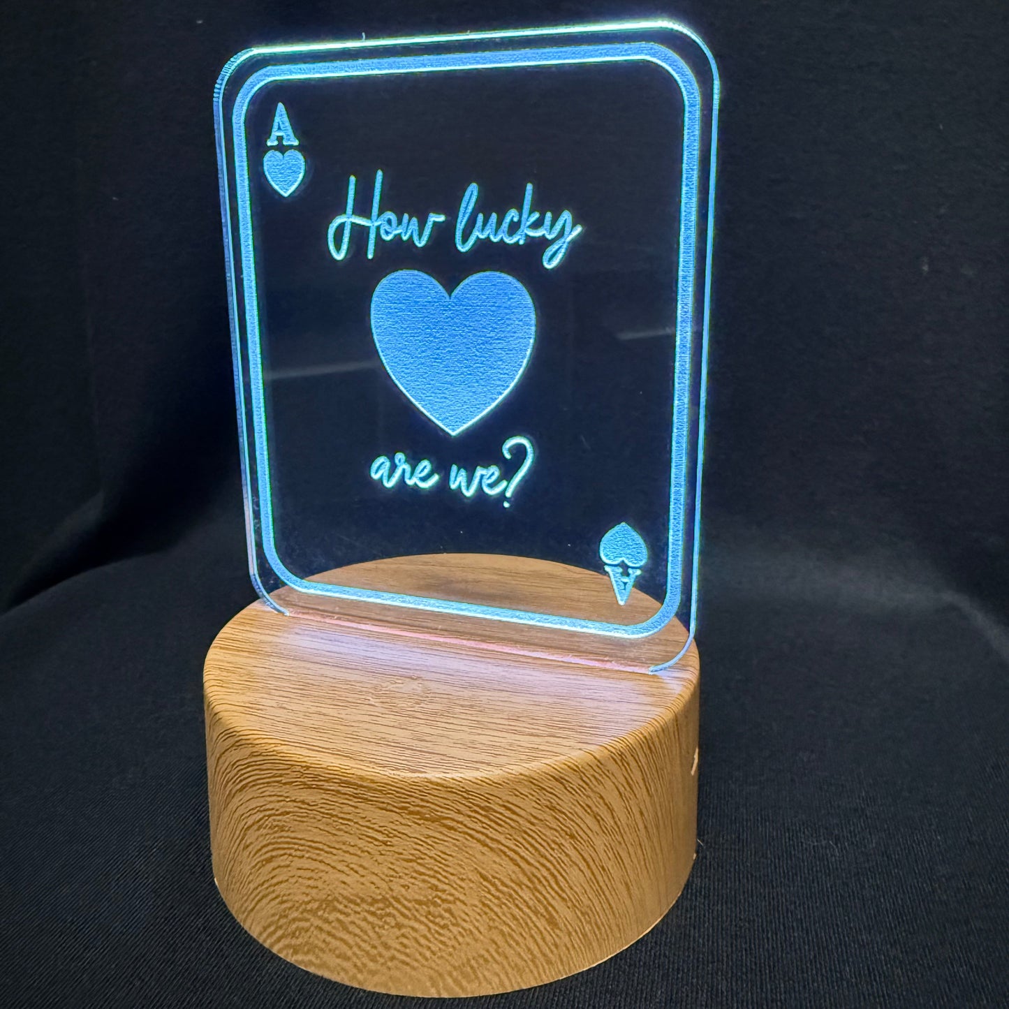 An LED acrylic light sign shaped like an Ace of Hearts playing card, displaying the text 'How lucky are we?' with a glowing blue heart, mounted on a cylindrical wooden base against a black background.