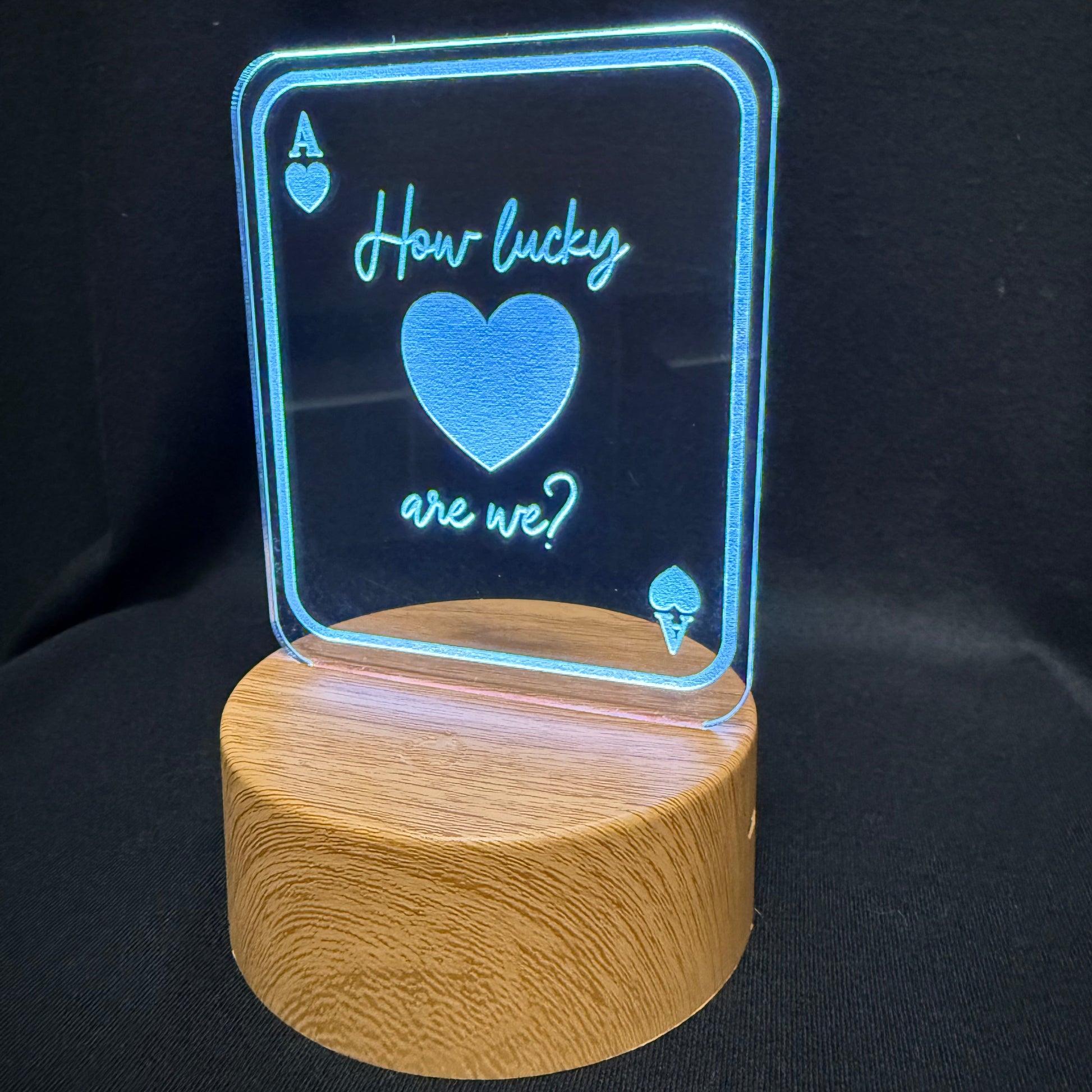 An LED acrylic light sign shaped like an Ace of Hearts playing card, displaying the text 'How lucky are we?' with a glowing blue heart, mounted on a cylindrical wooden base against a black background.