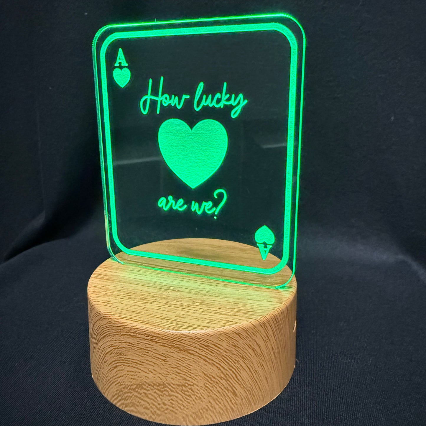 A wooden cylindrical base supporting a clear acrylic sign shaped like an ace of hearts card, glowing green with the text 'How lucky are we?' and a large heart, set against a dark fabric background.