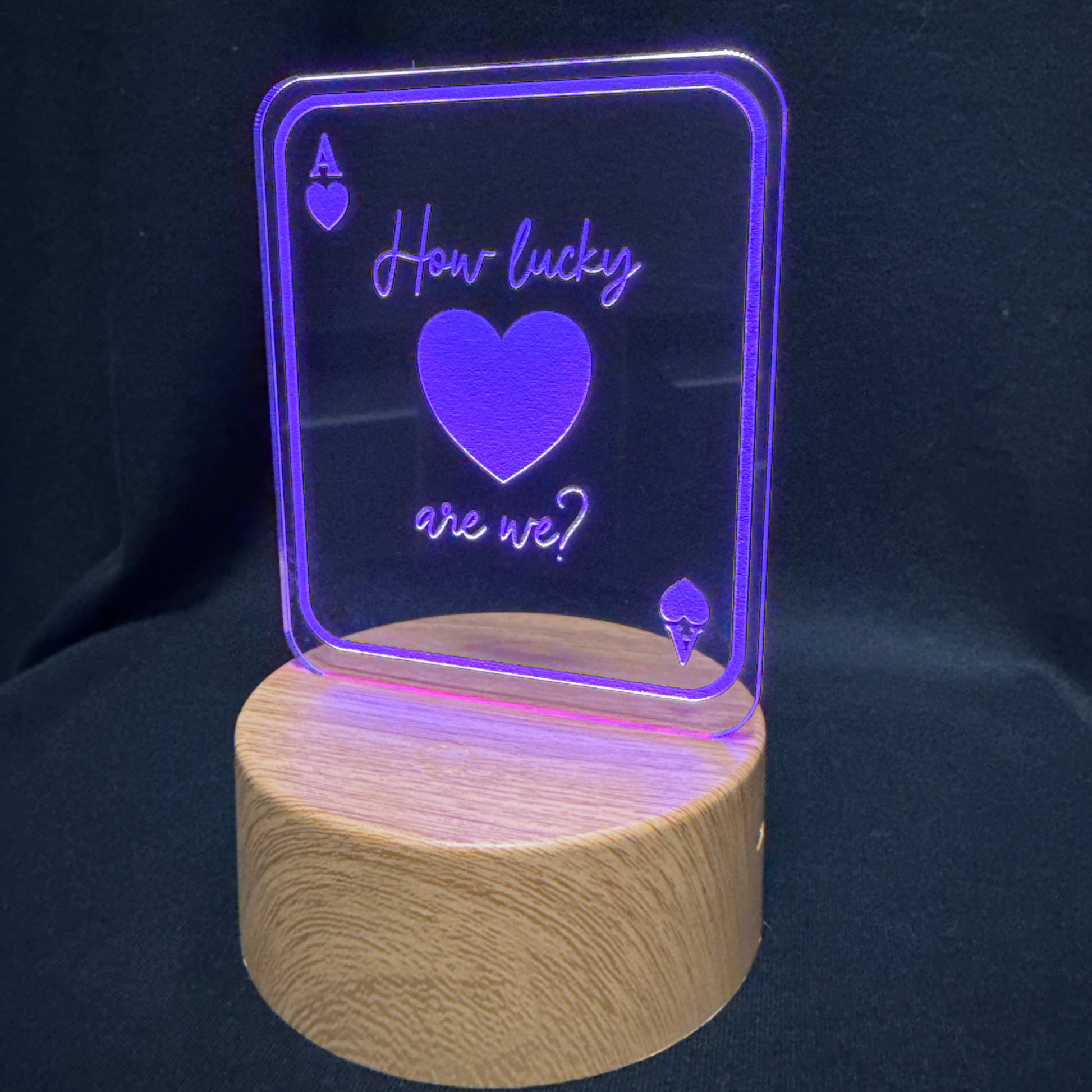 A wooden cylindrical lamp base supporting a clear acrylic sign with a purple LED glow. The sign features an Ace of Hearts symbol and the phrase 'How lucky are we?' in purple lettering, set against a dark background.
