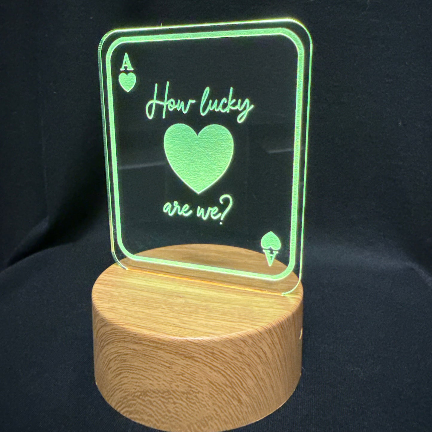 A wooden cylindrical lamp base supporting a clear acrylic sign that glows green with the text 'How lucky are we?' and an Ace of hearts design, set against a black background.