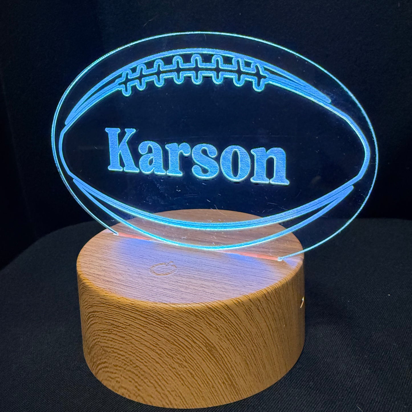 A personalized 3D optical illusion lamp featuring an illuminated football-shaped acrylic design with the name 'Karson' engraved, resting on a cylindrical wooden base against a black background.