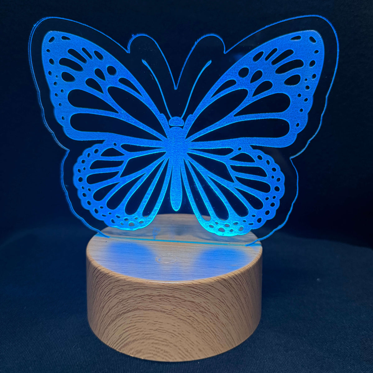 A butterfly-shaped LED night light with a glowing blue acrylic butterfly design mounted on a round wooden base, set against a dark blue background.