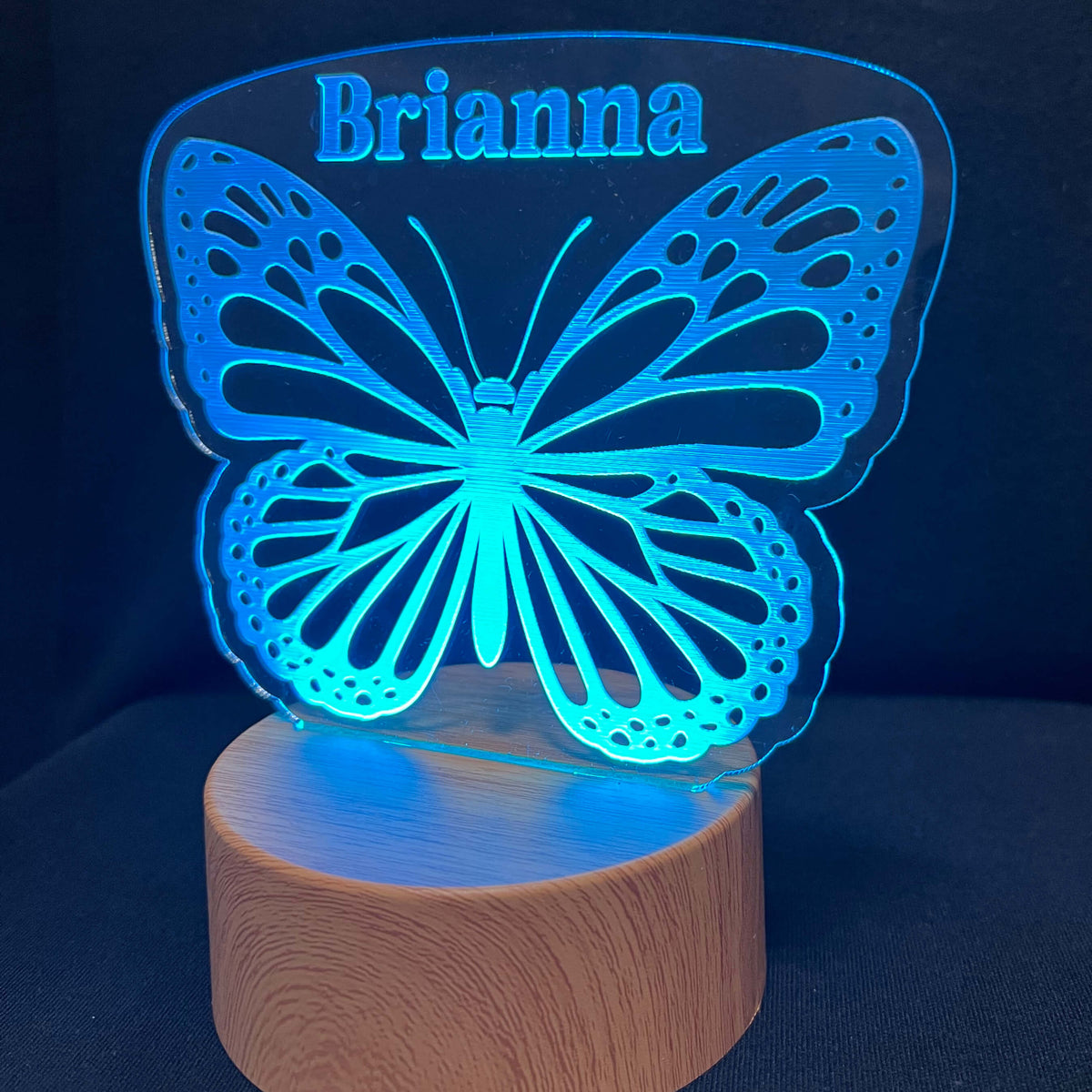 A personalized LED night light featuring a glowing blue butterfly silhouette with the name 'Brianna' cut out at the top, resting on a cylindrical wooden base against a dark background.