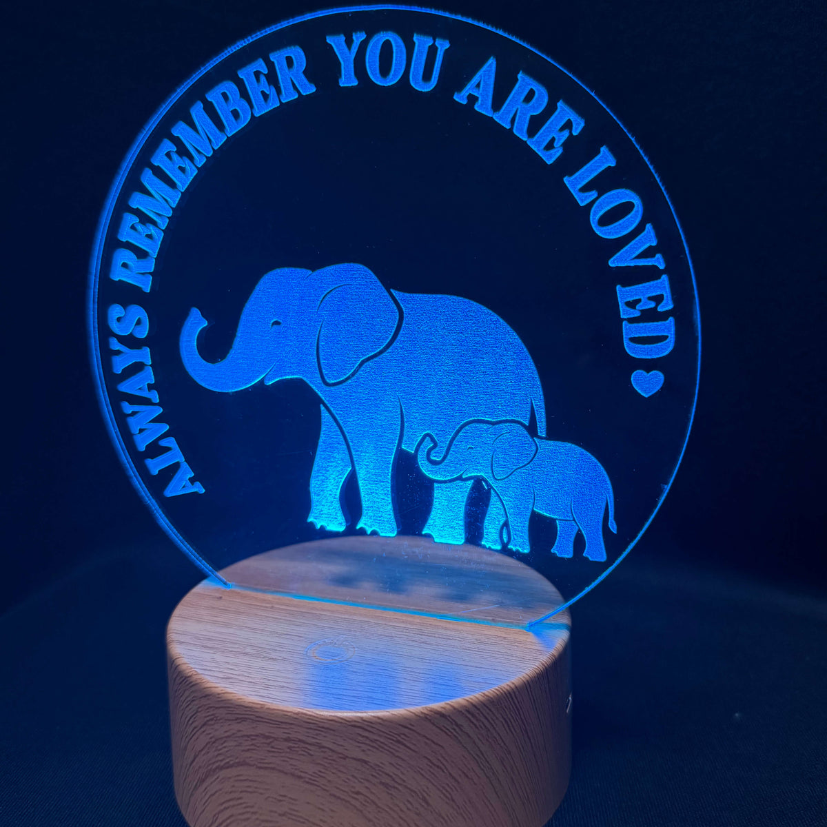 A 3D acrylic LED night light featuring a glowing blue silhouette of a mother and baby elephant with the text 'ALWAYS REMEMBER YOU ARE LOVED' in a circular design, mounted on a cylindrical wooden base.