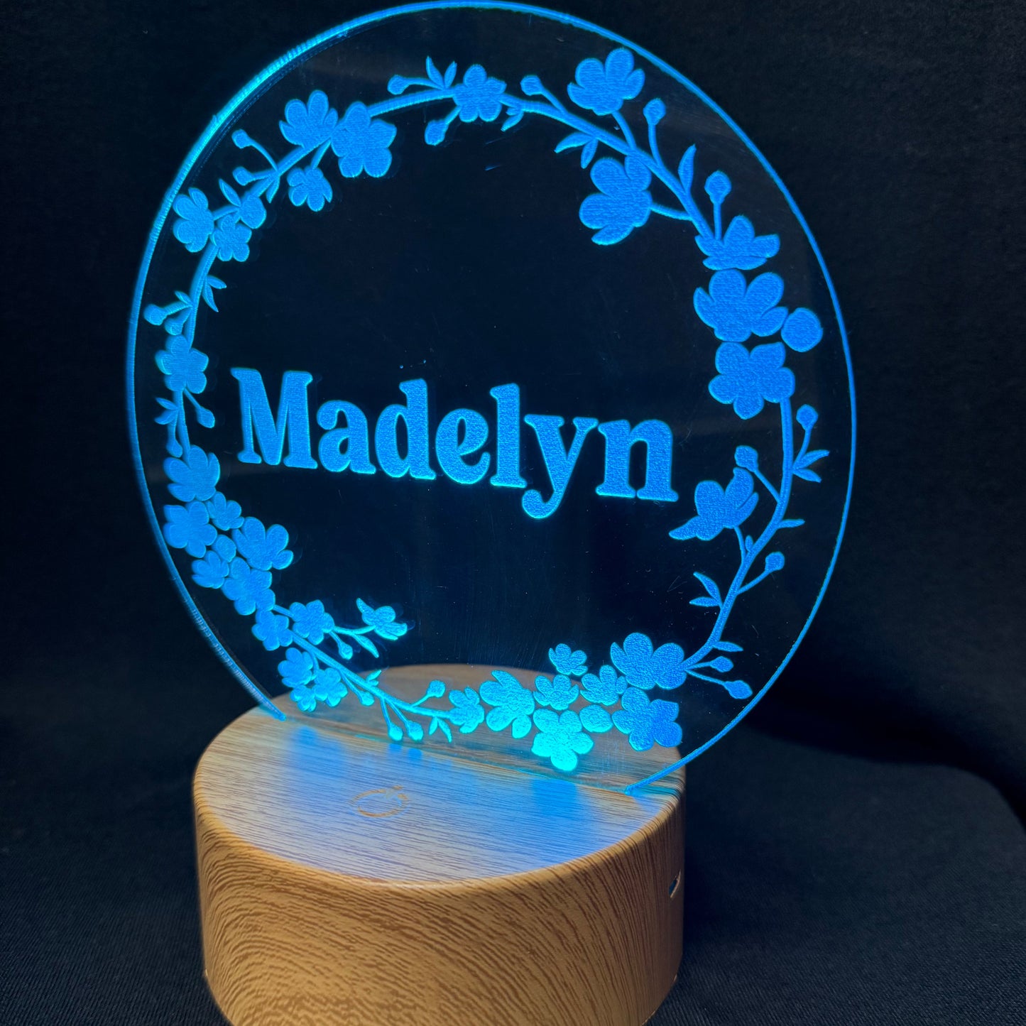 A personalized acrylic LED night light featuring the name 'Madelyn' in blue lettering, surrounded by a floral wreath design, mounted on a round wooden base against a black background.