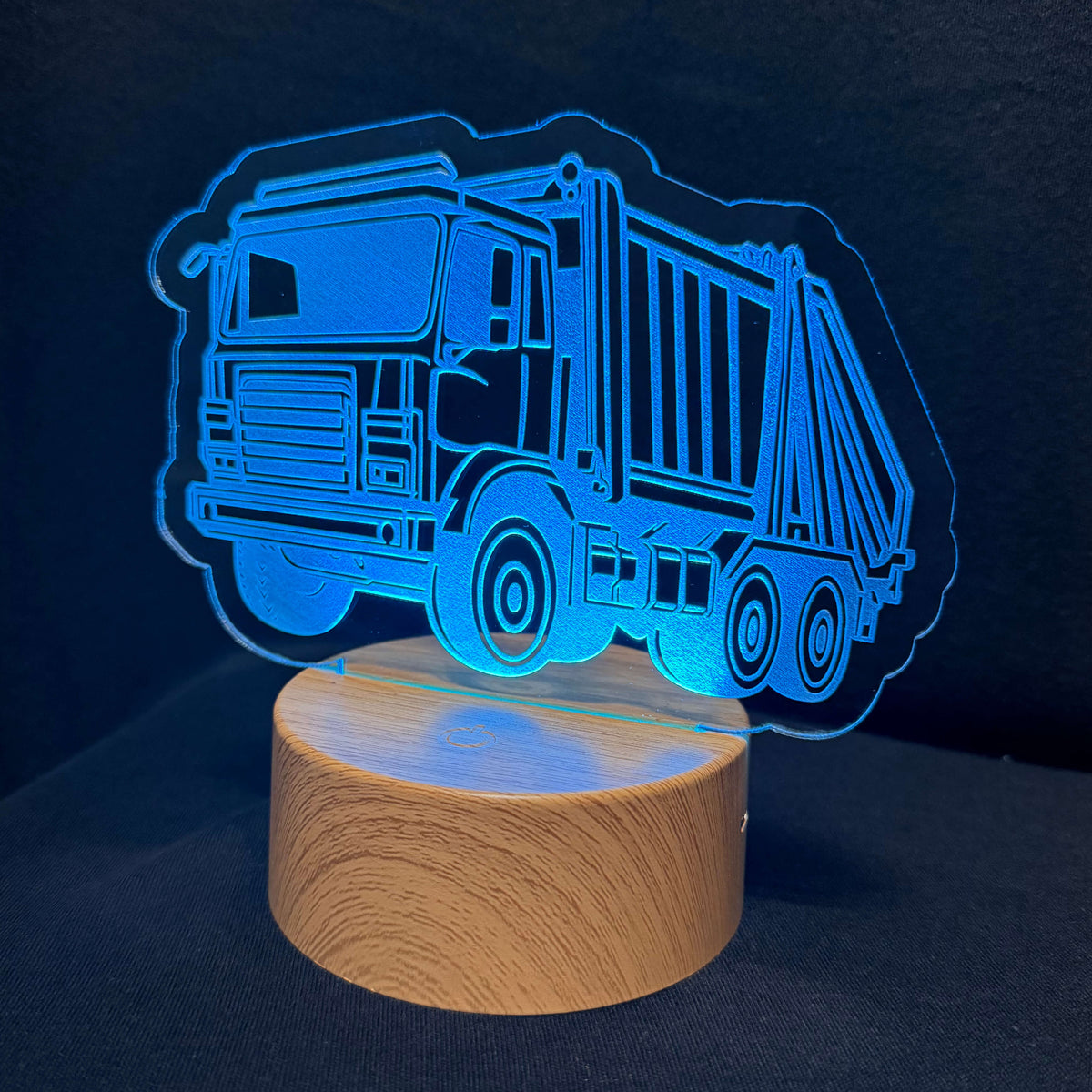 A truck-shaped LED night light with a glowing blue acrylic cutout and a round wooden base, shown on a dark background.