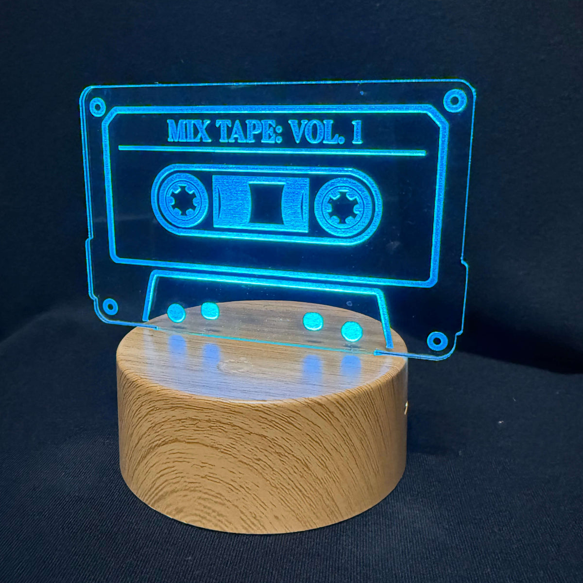A glowing acrylic cassette tape lamp with the text 'MIX TAPE: VOL. 1' on a round wooden base, set against a dark black background.