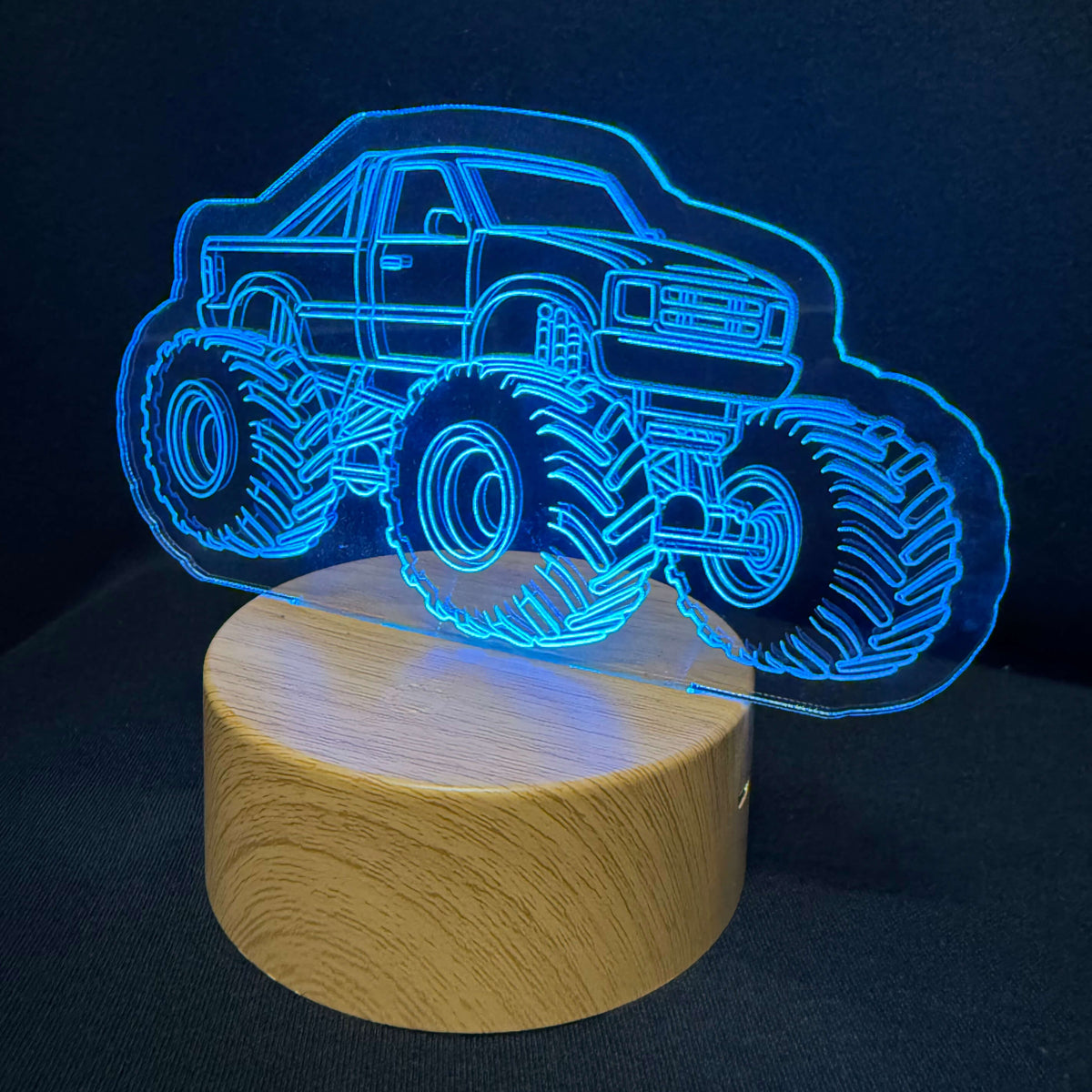 A 3D optical illusion lamp featuring a glowing blue monster truck outline on a round, light wood-grain base, set against a dark background.