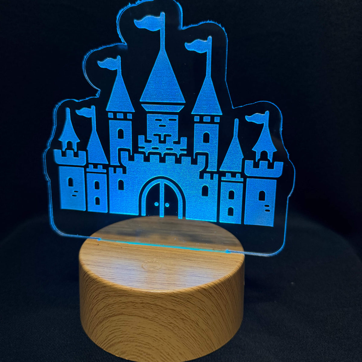 A blue illuminated acrylic castle-shaped night light on a round wooden base, photographed against a dark black background.