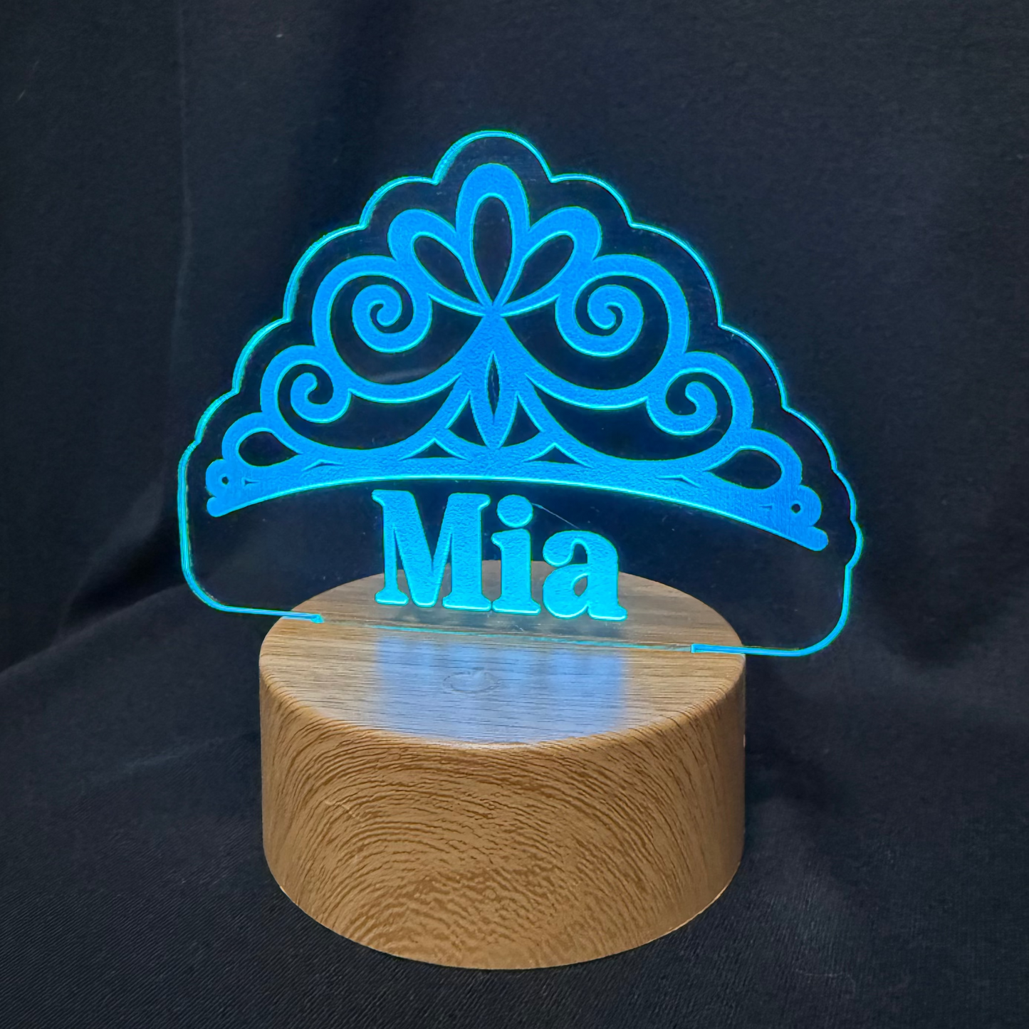 Blue tiara-shaped light with 'Mia' on a wooden base against a dark background