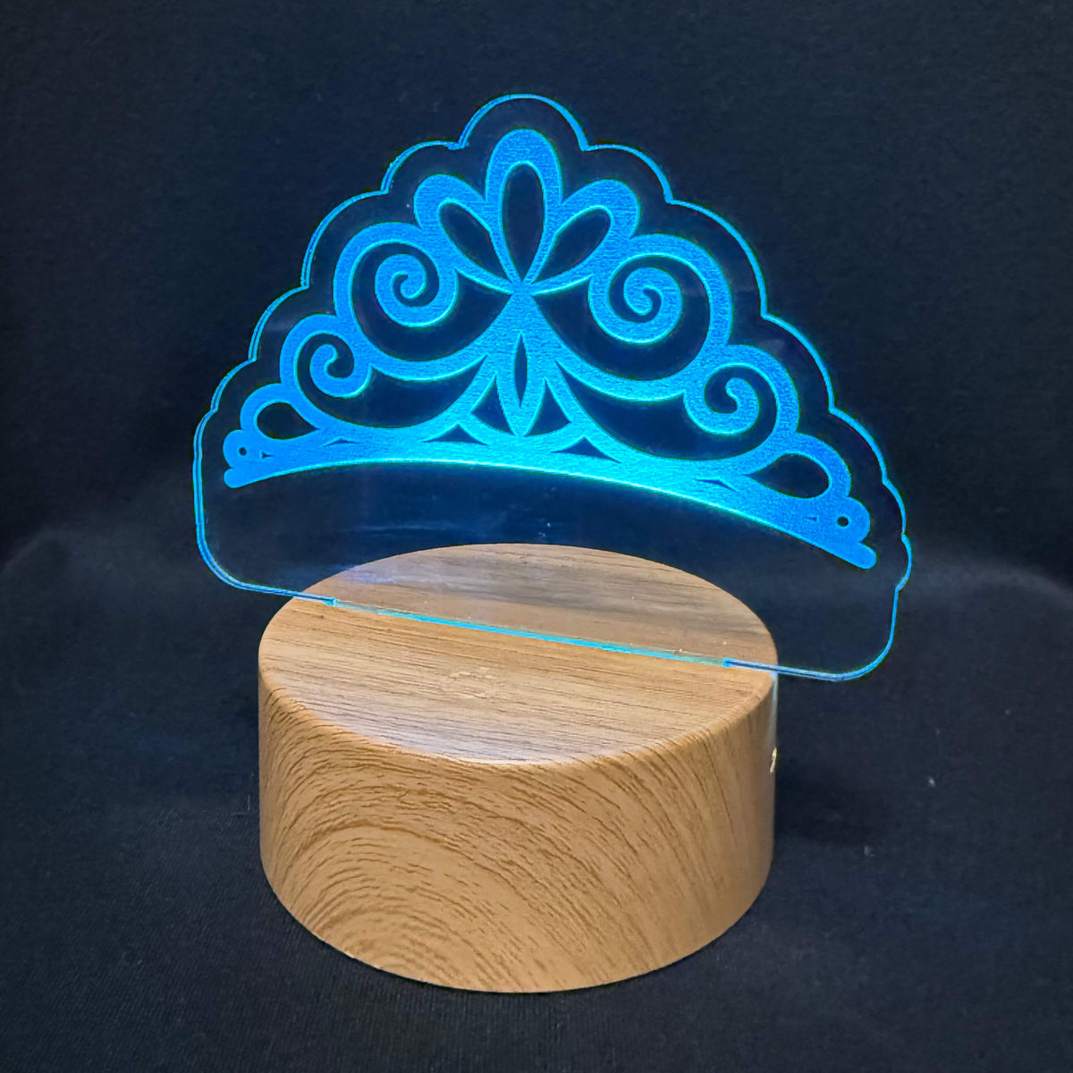A 3D optical illusion night light featuring a glowing blue tiara design projected above a cylindrical wooden base, set against a dark black fabric background.