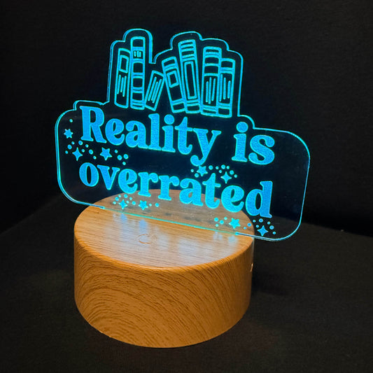 A 3D acrylic LED night light with the text 'Reality is overrated' in glowing cyan blue, mounted on a cylindrical wooden base and photographed against a dark background.