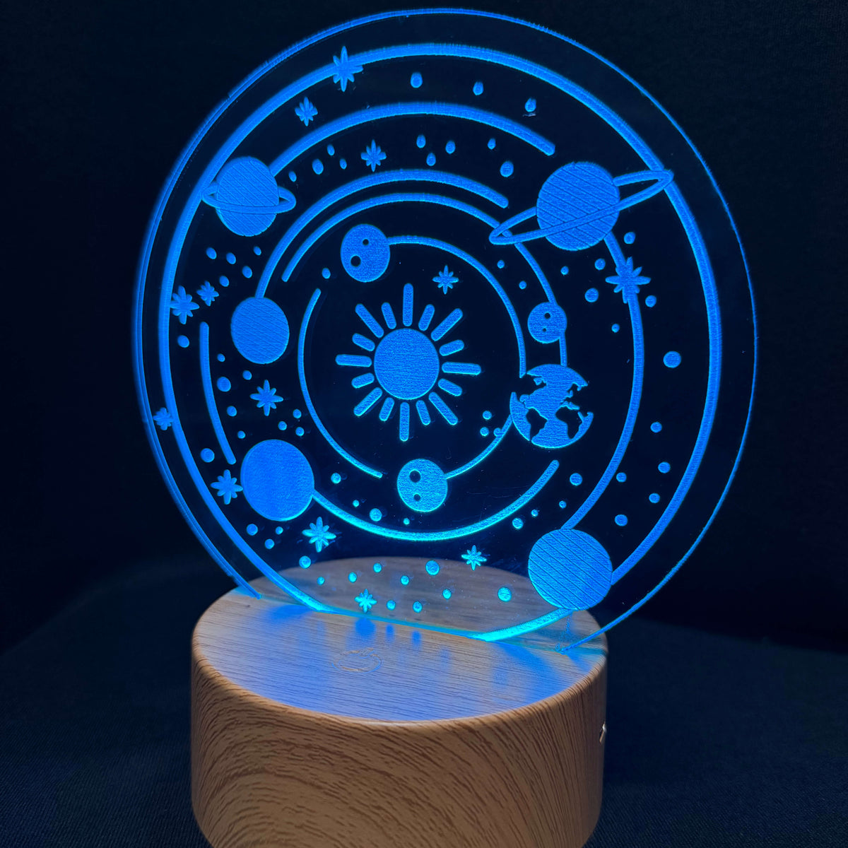An LED acrylic night light featuring a glowing blue solar system design with planets, stars, and a sun, mounted on a round wooden base against a dark background.