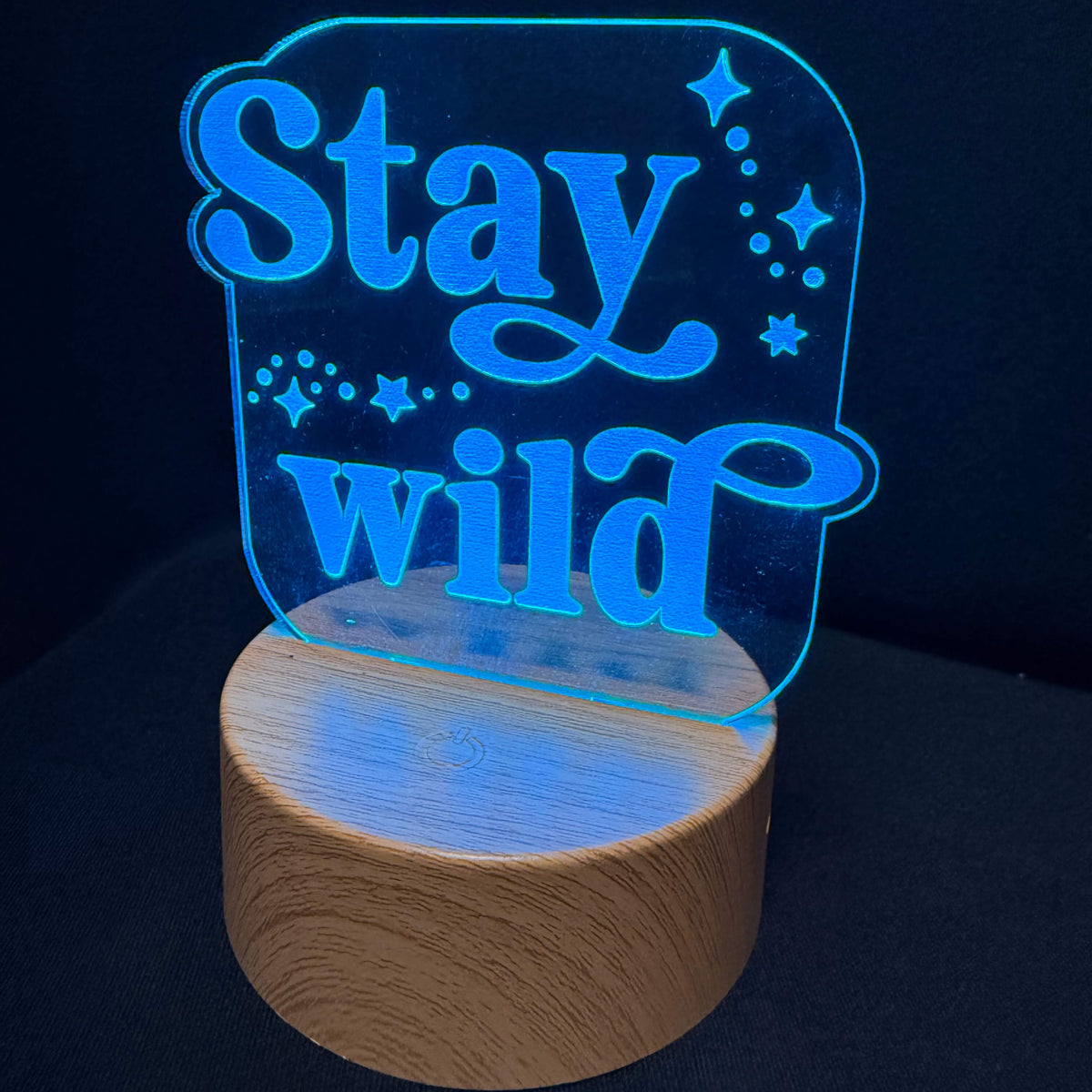 A modern LED acrylic night light with the phrase 'Stay wild' in glowing blue script, mounted on a round natural wood base against a dark background.