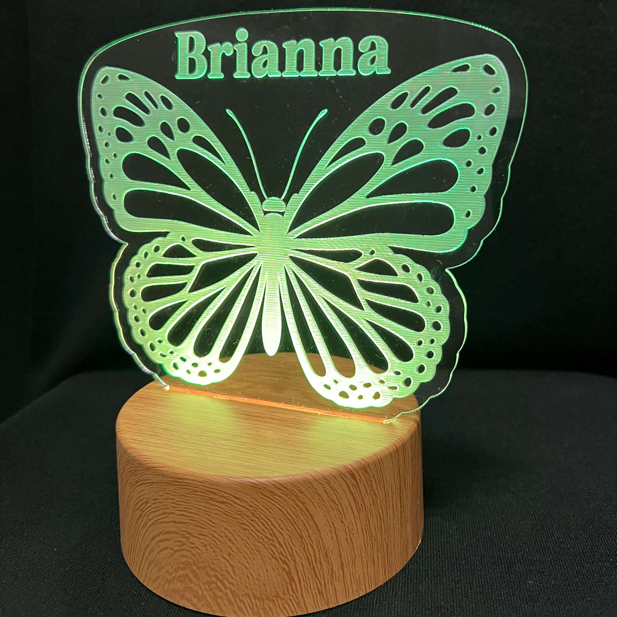 A personalized LED butterfly night light with the name 'Brianna' cut into the top wing, glowing green and resting on a cylindrical wooden base against a black background.