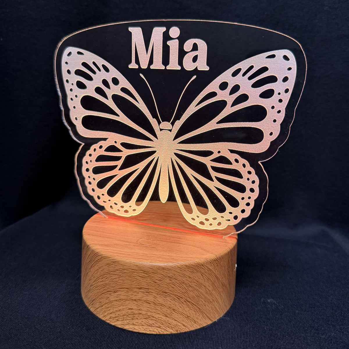 A personalized LED acrylic night light featuring a butterfly design and the name 'Mia' cut out at the top, mounted on a round wooden base against a dark blue background.