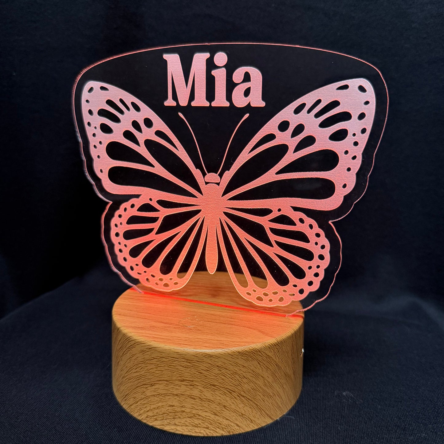 A personalized LED acrylic night light featuring a butterfly design with the name 'Mia' cut out in the center, mounted on a round wooden base against a dark fabric background.