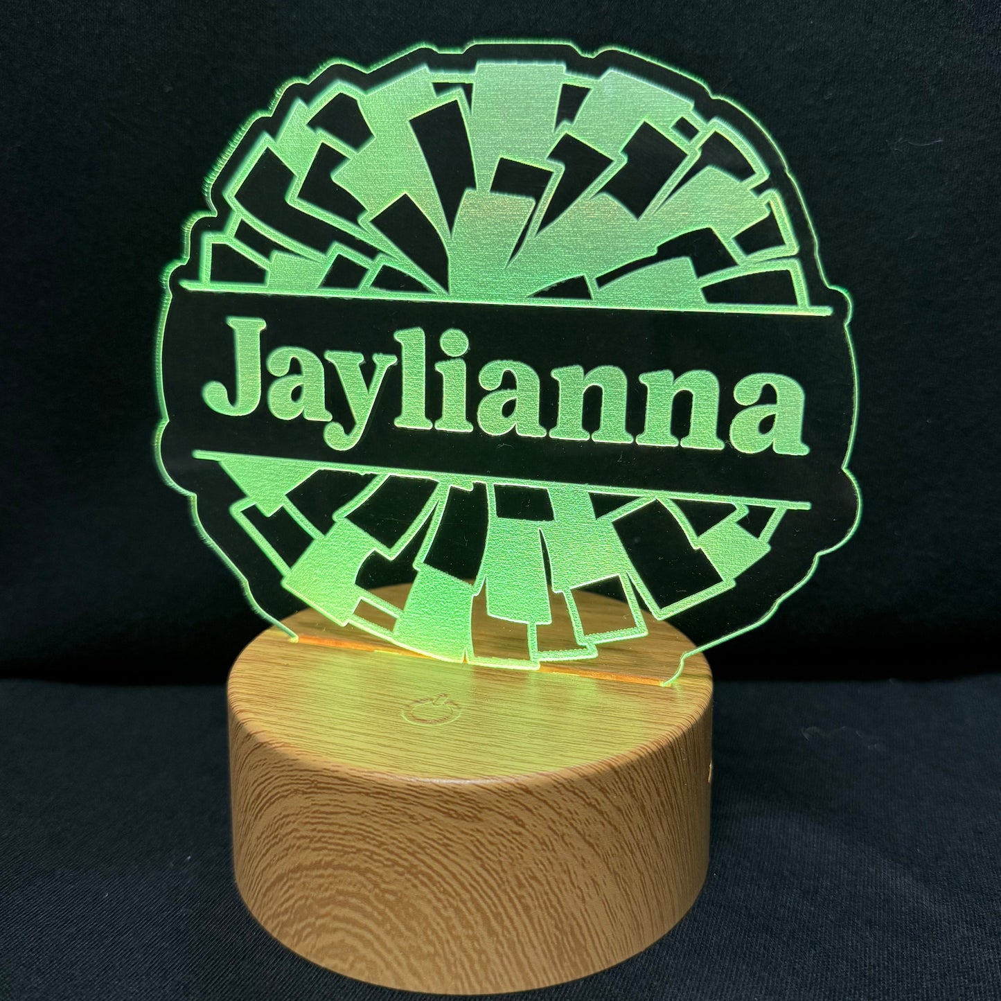 A personalized LED acrylic lamp featuring the name 'Jaylianna' in green on a black background, mounted on a round wooden base and photographed against a dark backdrop.