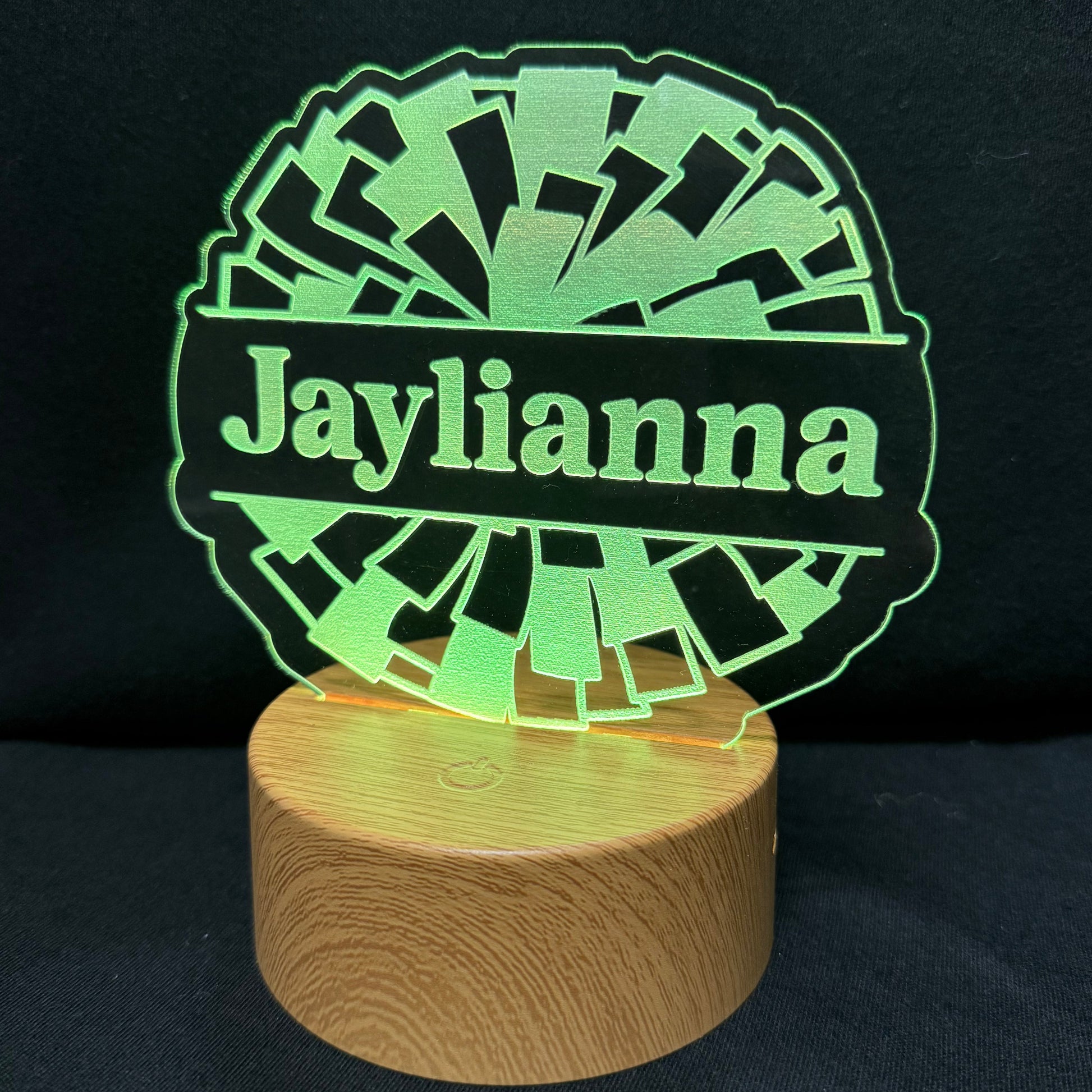 A personalized LED acrylic lamp featuring the name 'Jaylianna' in green on a black background, mounted on a round wooden base and photographed against a dark backdrop.