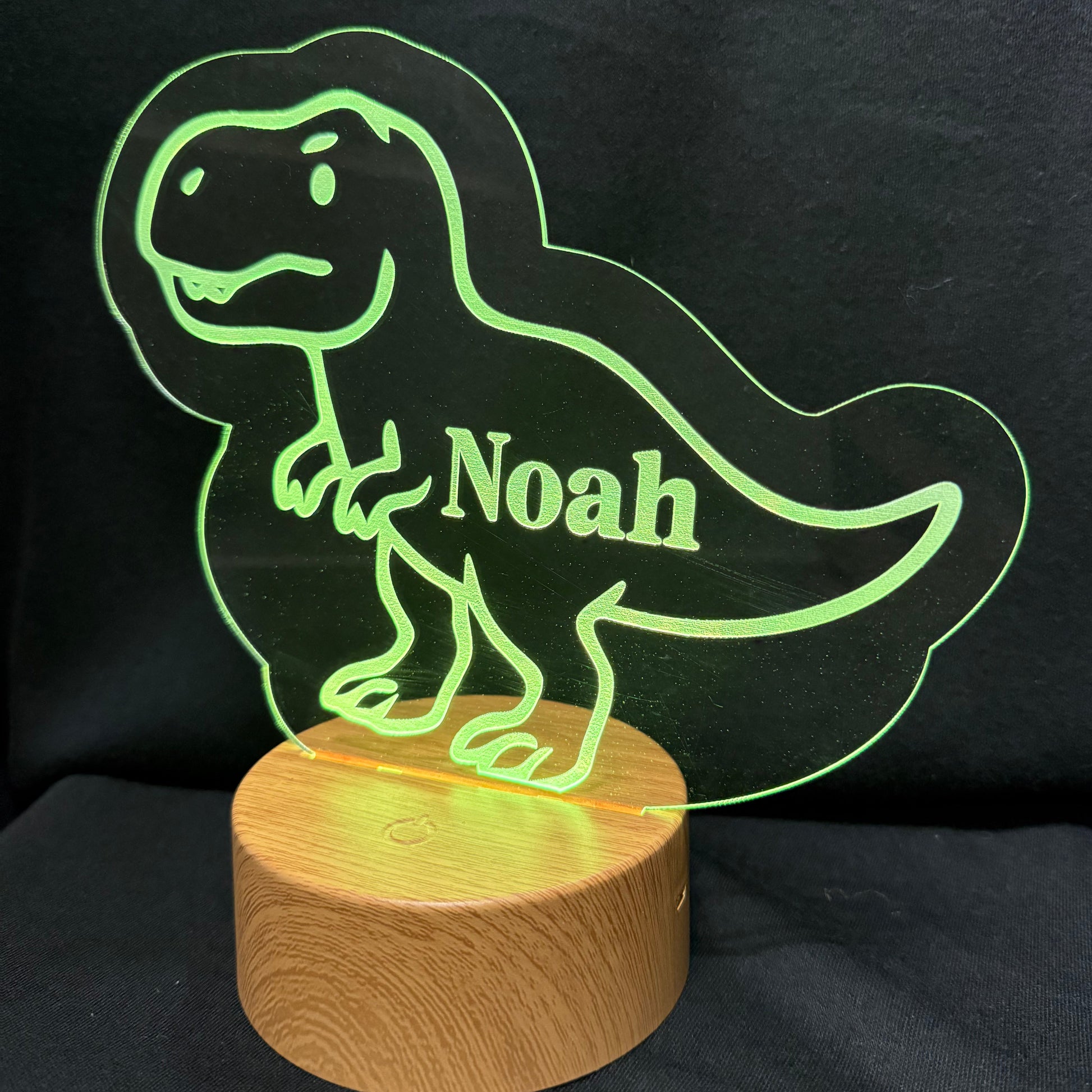A personalized LED night light shaped like a cartoon T-rex dinosaur, glowing green with the name 'Noah' cut out in the center, mounted on a round wooden base against a black fabric background.