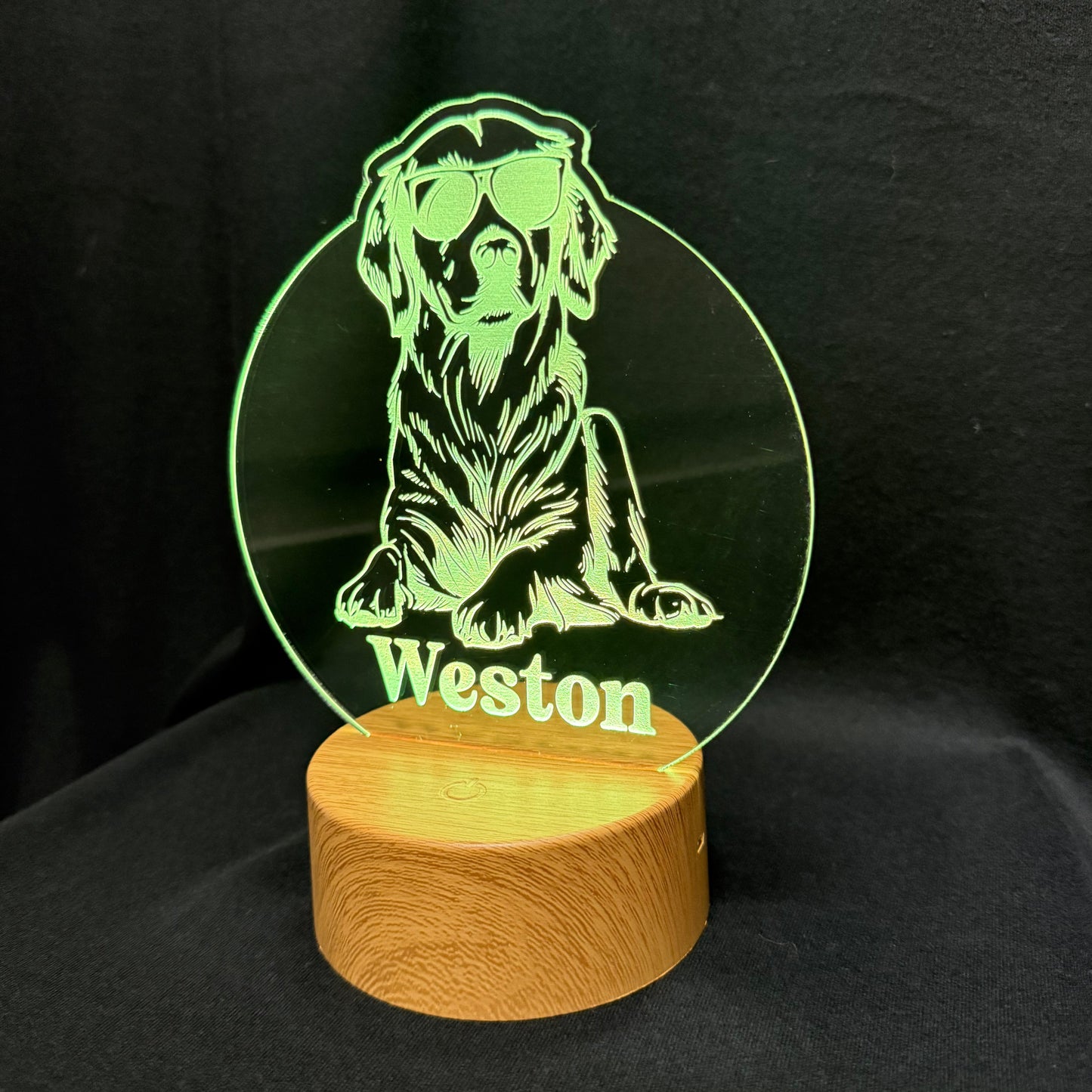 A personalized acrylic LED lamp featuring a laser-engraved green silhouette of a dog wearing sunglasses with the name 'Weston' below it, mounted on a round wooden base against a black background.