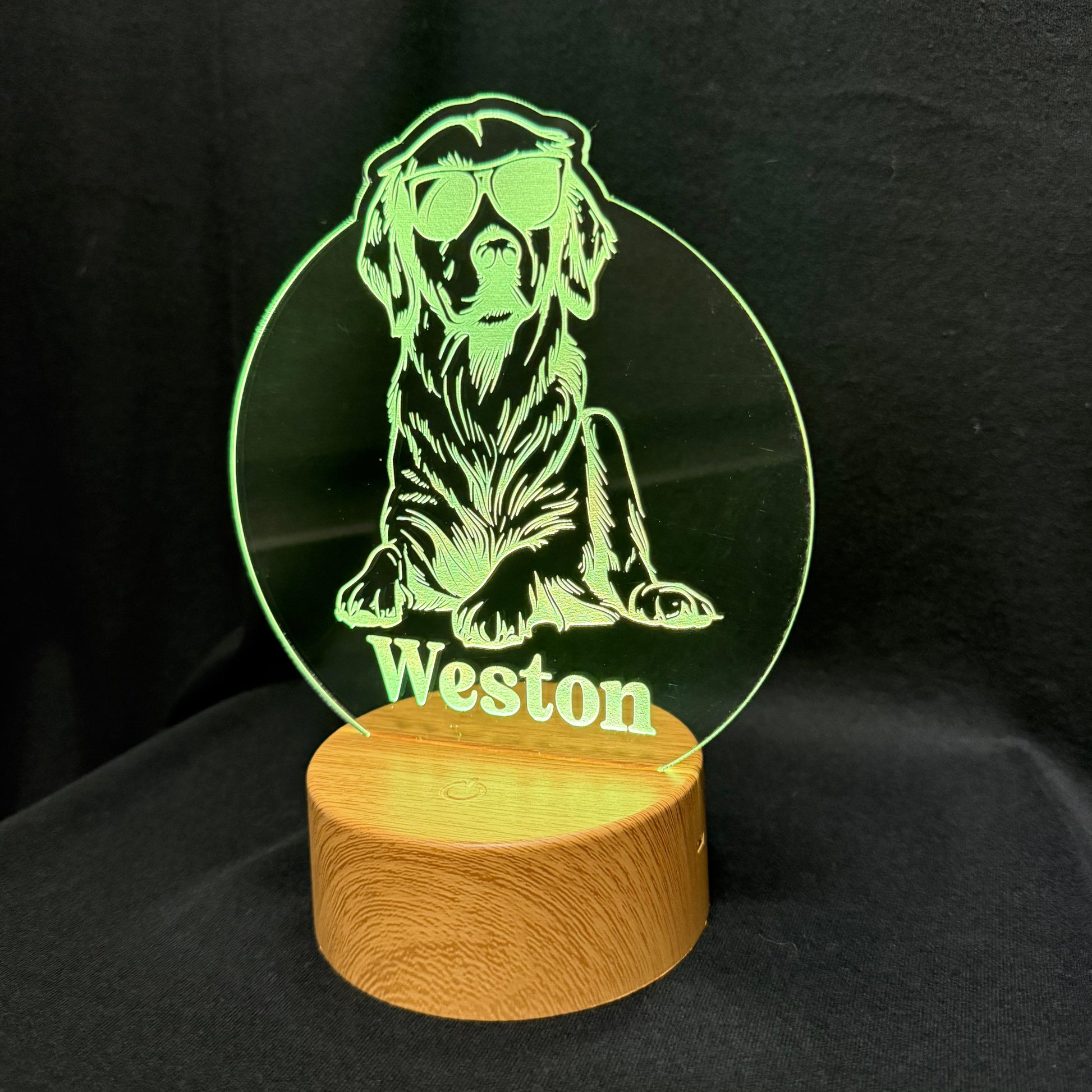 A personalized acrylic LED lamp featuring a laser-engraved green silhouette of a dog wearing sunglasses with the name 'Weston' below it, mounted on a round wooden base against a black background.