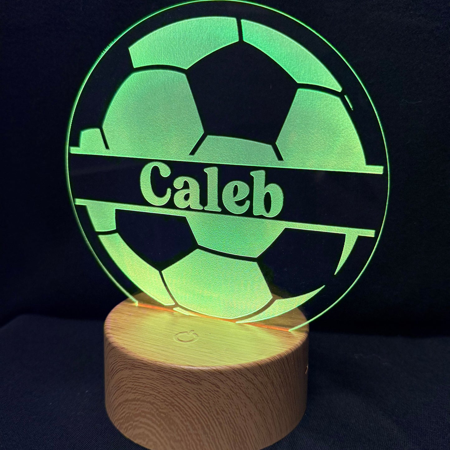 A personalized LED light lamp featuring a soccer ball design with the name 'Caleb' engraved in the center, resting on a cylindrical wooden base against a dark background.