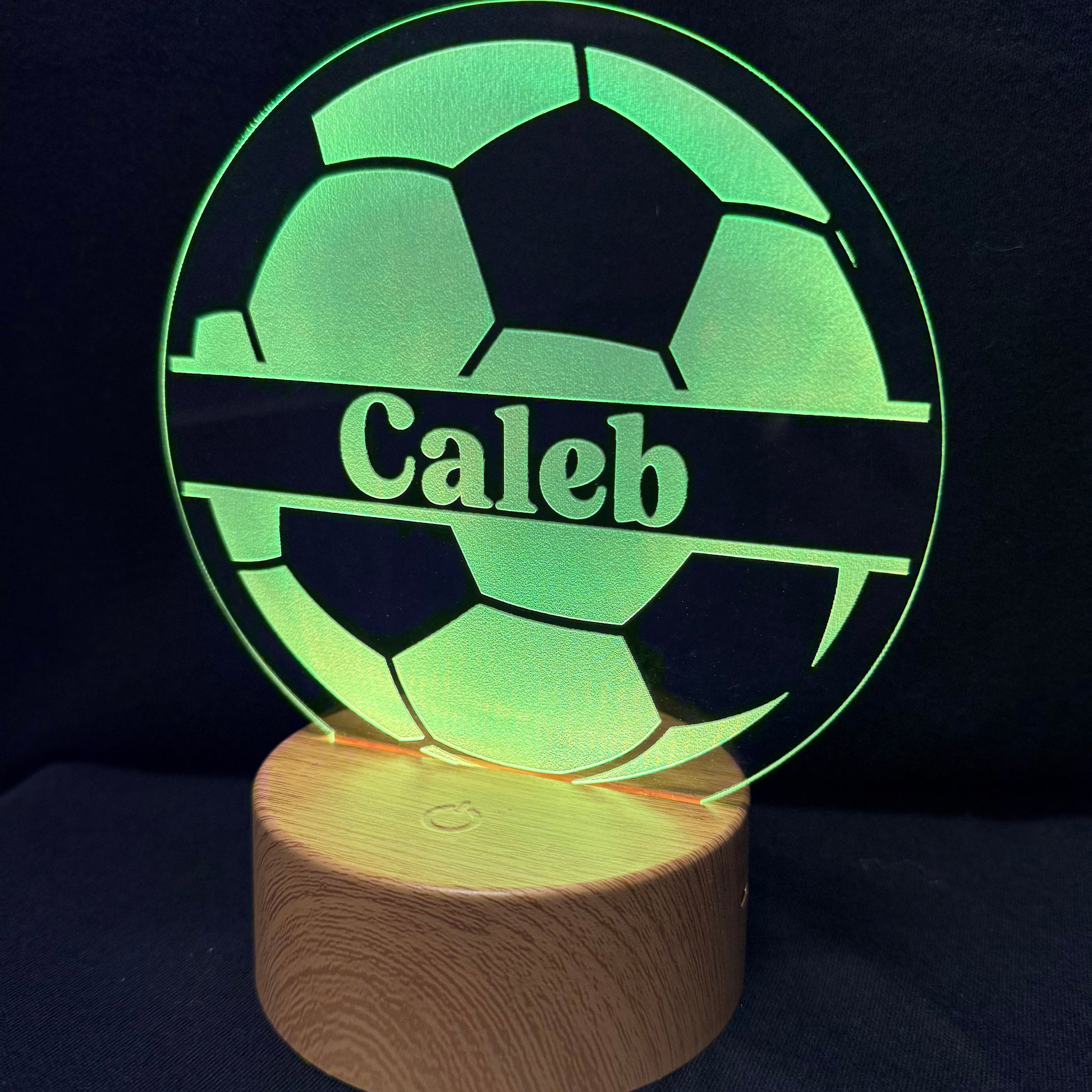 A personalized LED light lamp featuring a soccer ball design with the name 'Caleb' engraved in the center, resting on a cylindrical wooden base against a dark background.