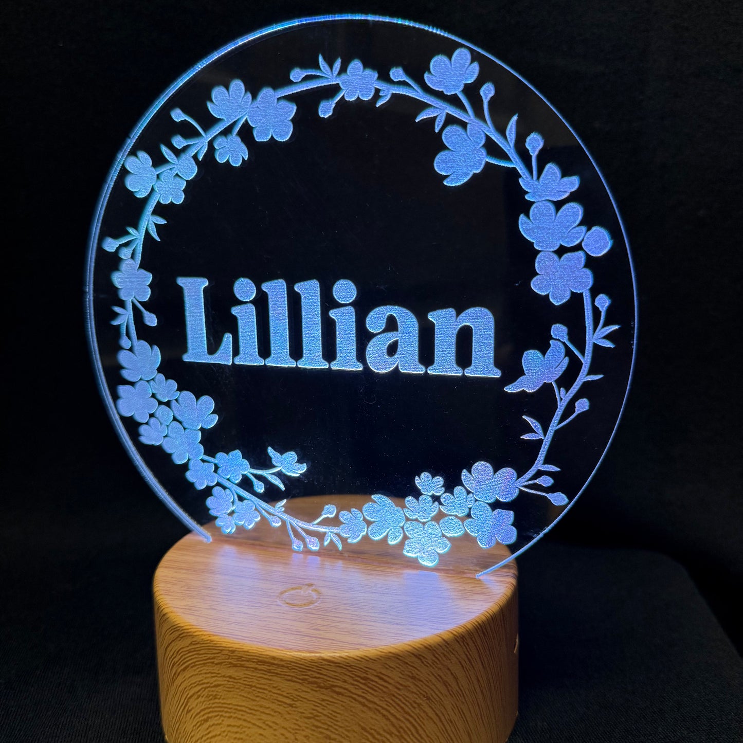 A personalized LED acrylic lamp featuring the name 'Lillian' in the center, surrounded by a glowing blue floral wreath, mounted on a cylindrical wooden base against a black background.