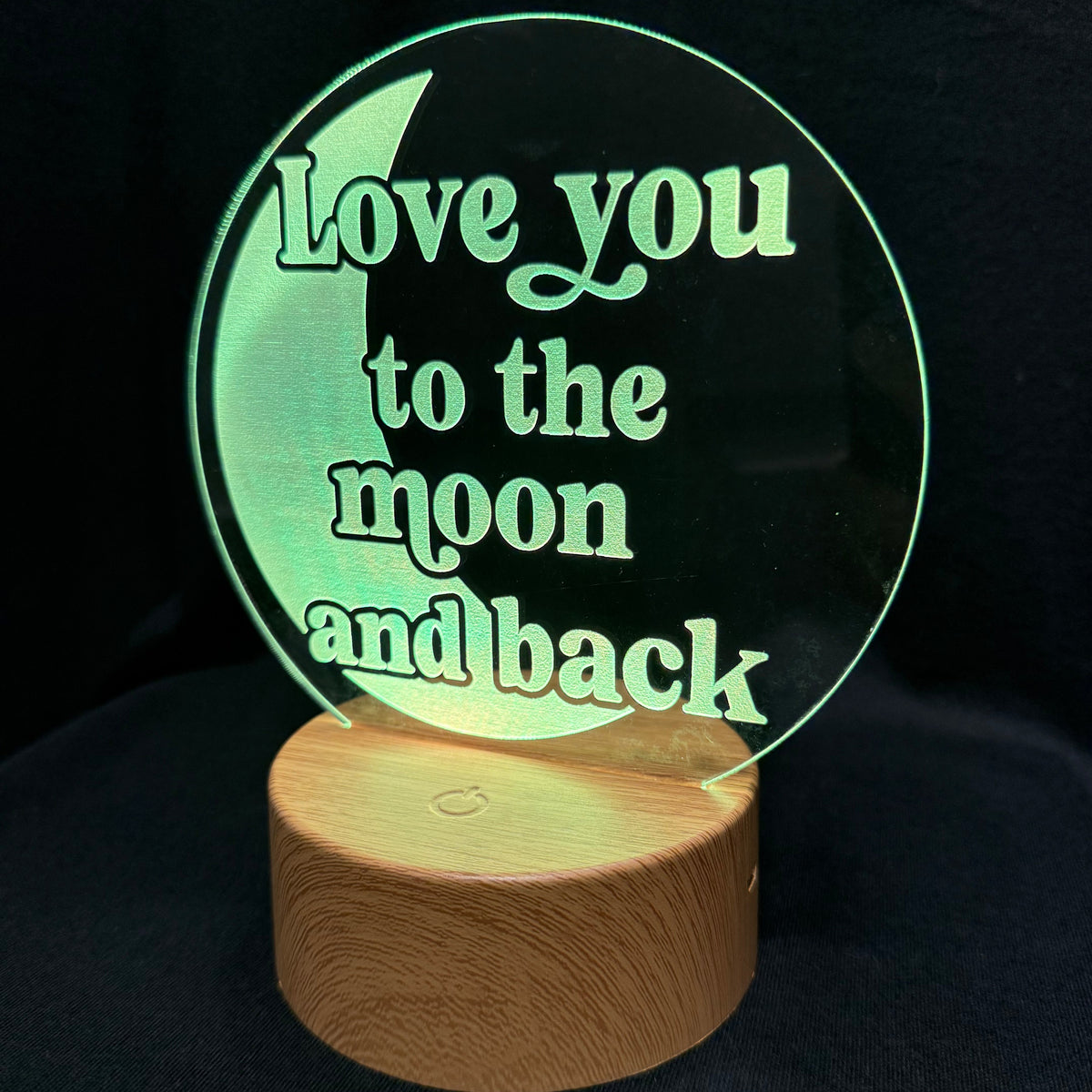 A circular acrylic LED night light with the phrase 'Love you to the moon and back' in light green lettering, mounted on a cylindrical wooden base against a black background.